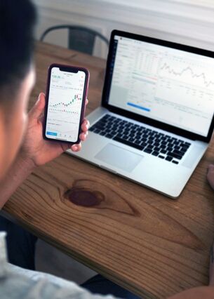 Ein Mann sitzt an einem Holztisch und betrachtet Finanz- und Börsendiagramme auf seinem Smartphone und Laptop. Das Bild zeigt eine moderne Arbeitssituation im Bereich Trading oder Finanzanalyse