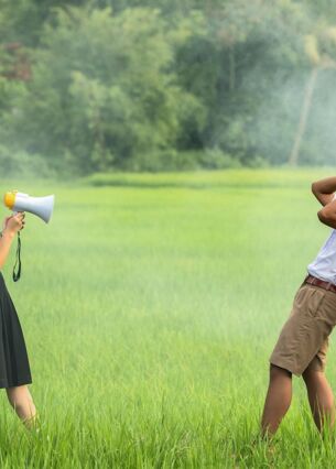 Eine Schülerin steht in einem grünen Feld und spricht durch ein Megafon, während zwei Schüler in Uniform sich die Ohren zuhalten und zurückweichen. Die Szene wirkt humorvoll und zeigt eine lebhafte Kommunikation im Freien