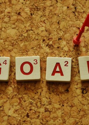 Das Wort ‚GOAL‘ ist aus Scrabble-ähnlichen Buchstabensteinen auf einem Korkbrett gelegt. Daneben steckt eine kleine rote Flagge, die symbolisch ein Ziel oder eine erreichte Etappe markiert.