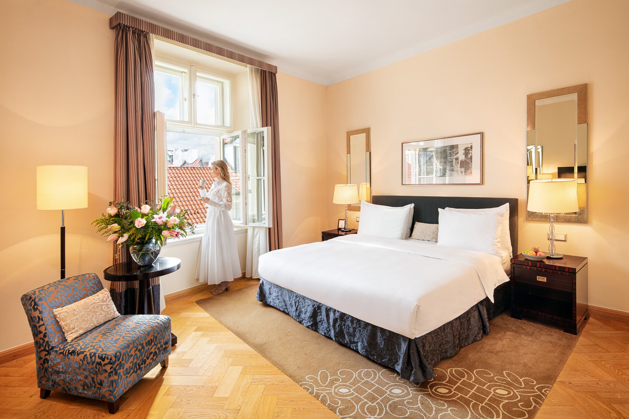 In a deluxe room with peach walls and golden wood floors, a white woman in a white dress drinks from a teacup and looks out open windows.