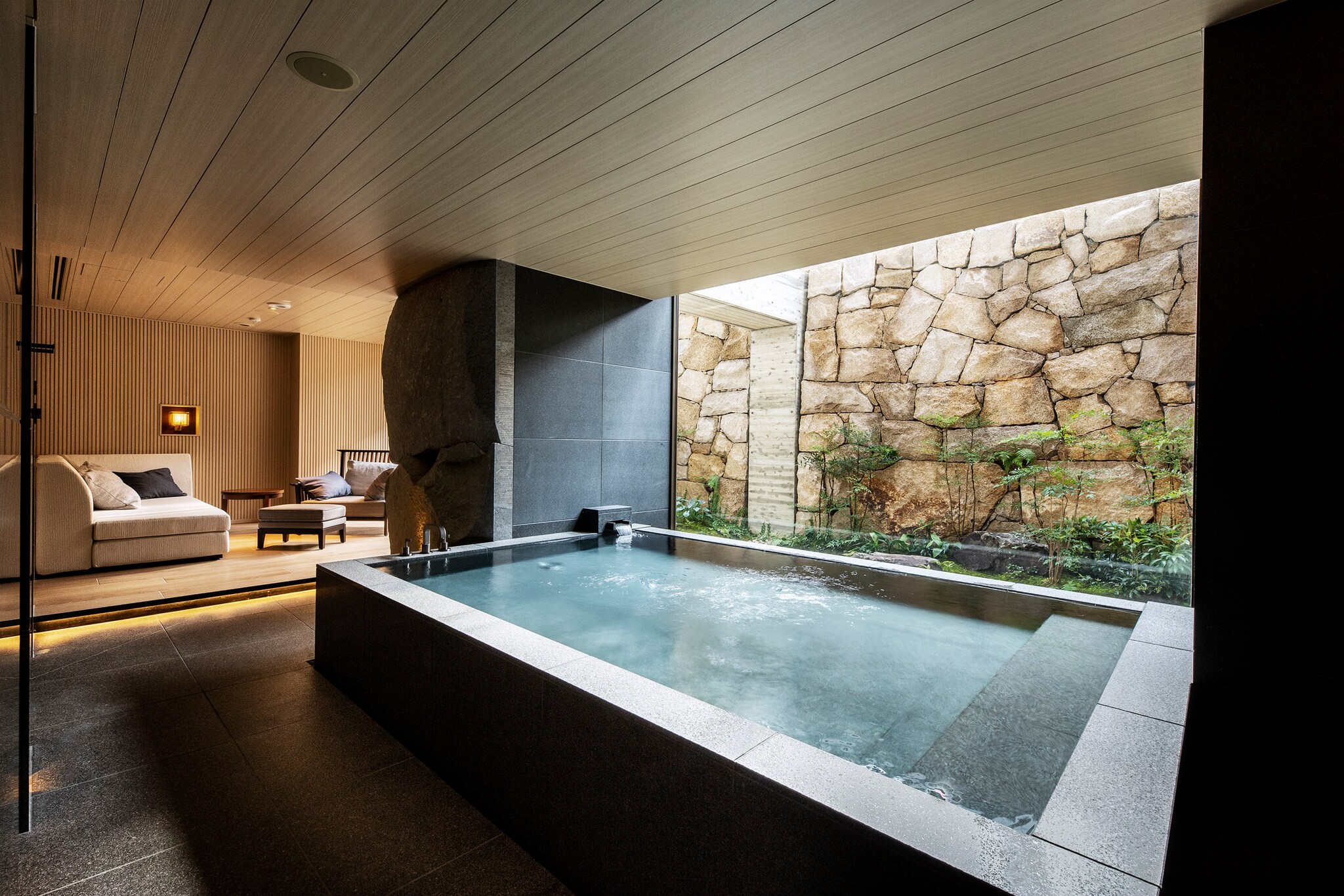 Interior of an Onsen Suite in which a rectangular hot tub sits burbling next to a light well lined with rocks and greenery.