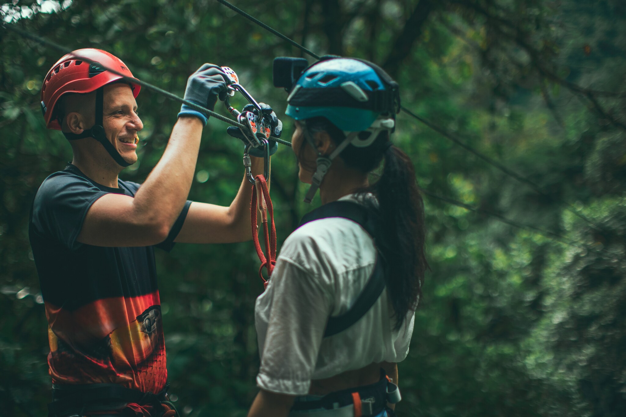 In a lush jungle, a white man in a red helmet affixes a carabiner to a zipline for an Asian woman wearing a blue helmet and safety harness.