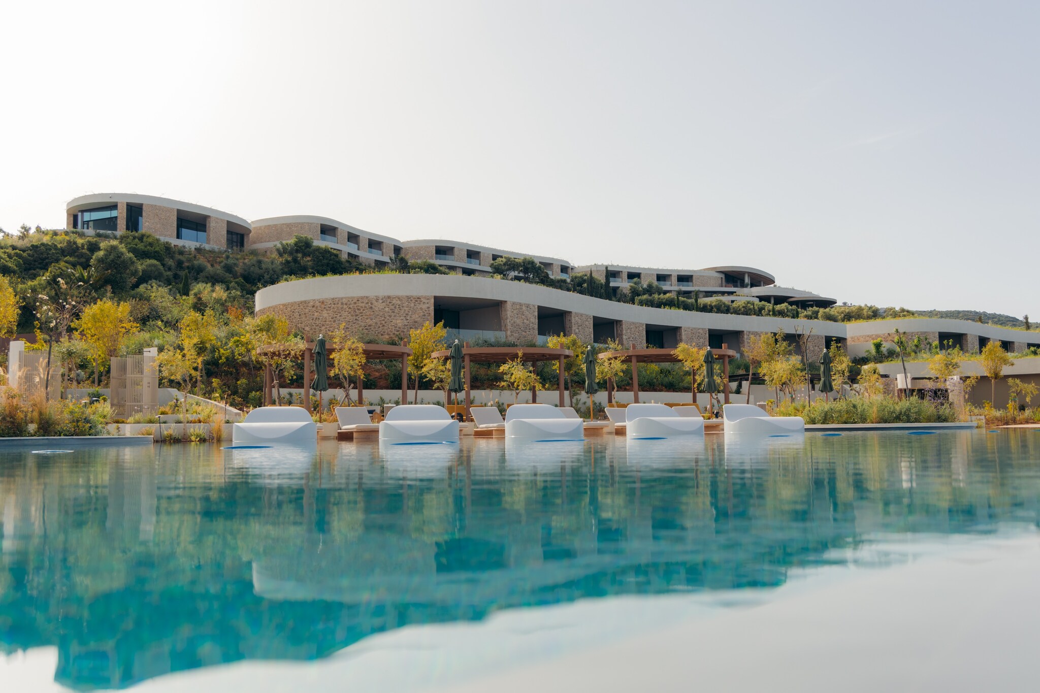 View over the surface of the hotel pool, at the hotel buildings set into the hillside and a set of large, curved white sun beds.