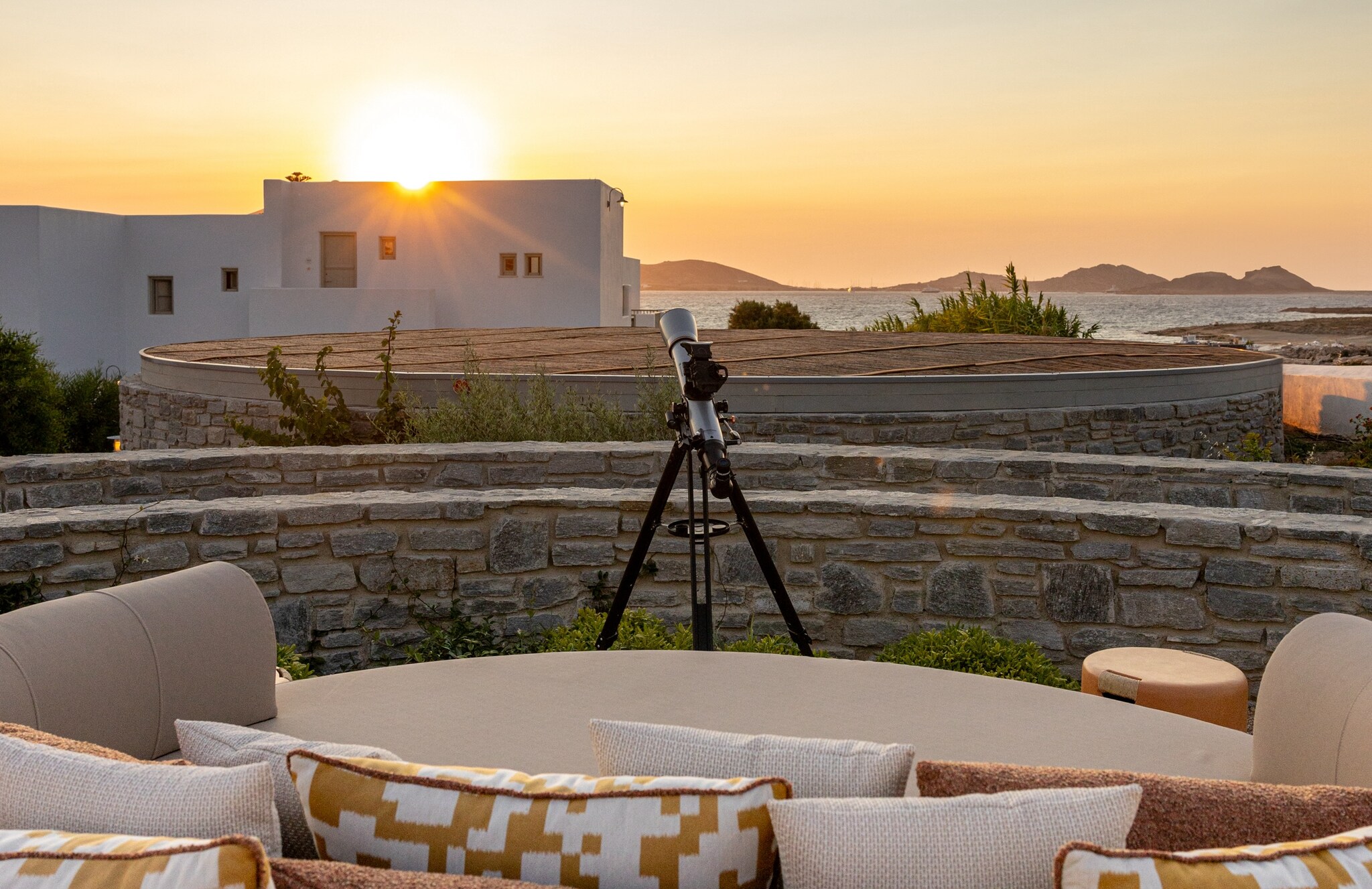 Beyond a curved tan outdoor couch, a telescope looks over low stone walls and white buildings at the setting sun.