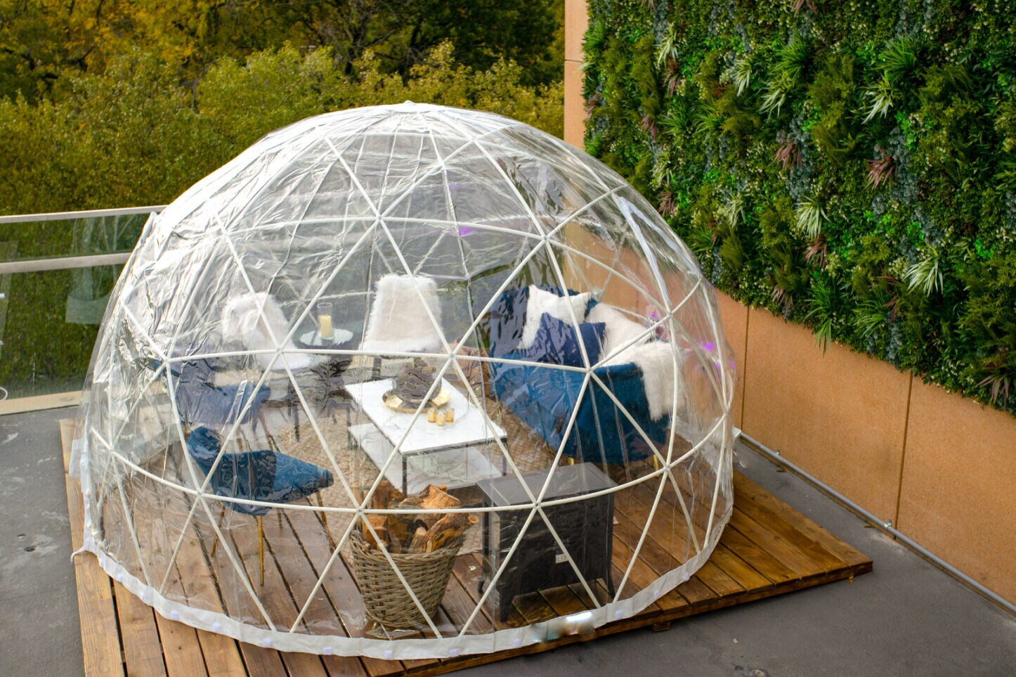 Exterior of a plastic geodesic dome on a wooden patio, containing a loveseat, several chairs, a portable fireplace, and a basket of wood.