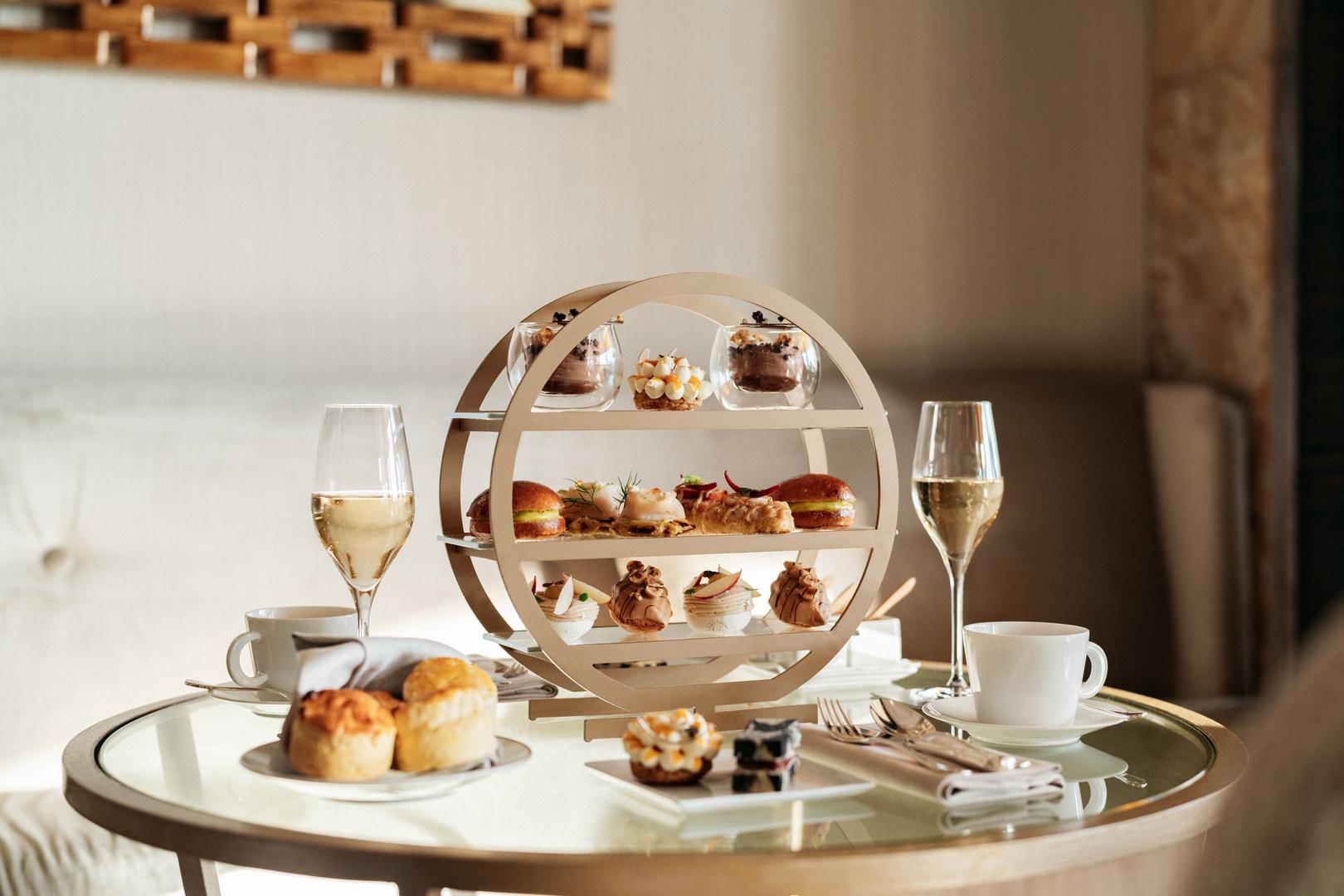A round glass table is elegantly set with a tiered tea tray of pastries, glasses of champagne, and mugs of tea.