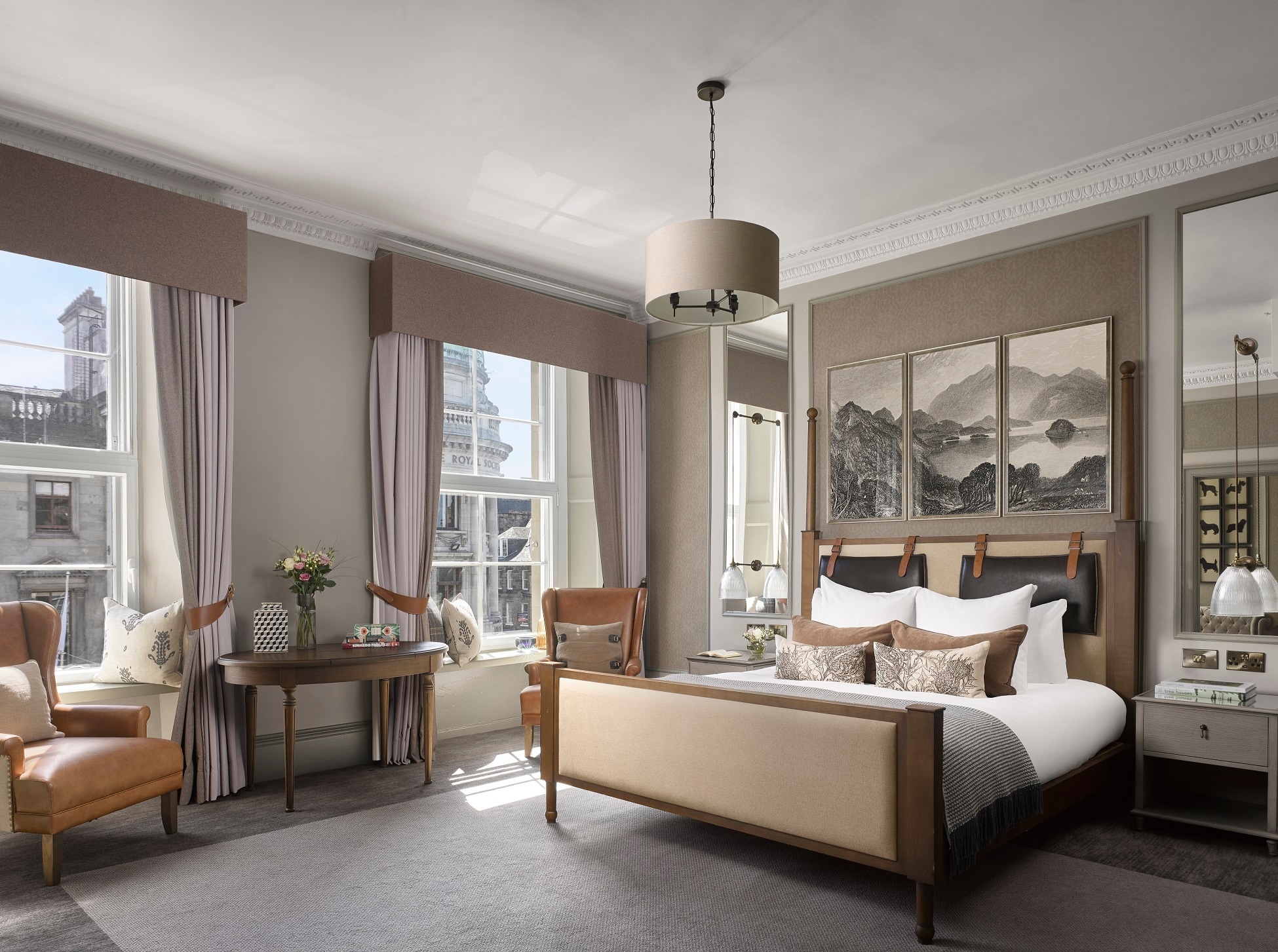 In a suite, a bed with a tan headboard sits under a triptych of black-and-white landscapes. Two window seats are framed by lilac curtains.