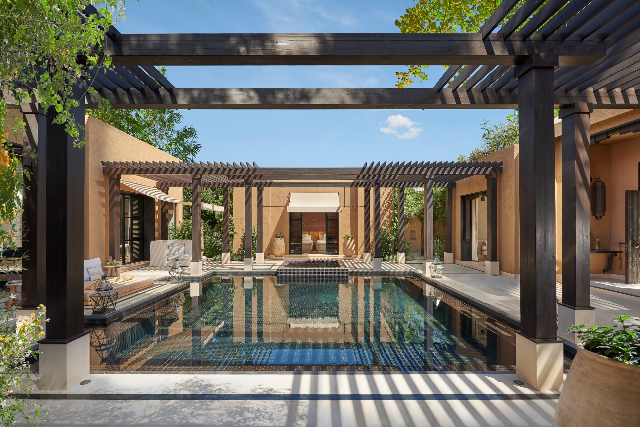 Outside of a villa, pergolas shade outdoor furniture and potted plants around a central square pool lined with small blue and green tiles.