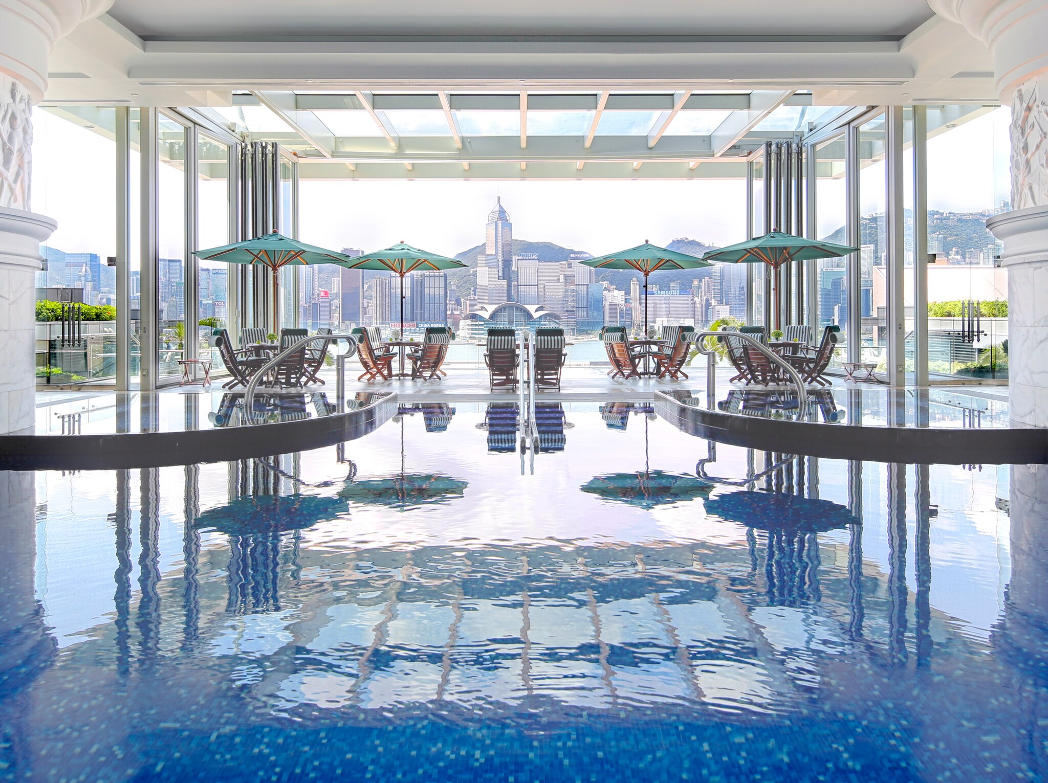 The indoor swimming pool is lined by small blue tiles. Beyond, a patio with umbrellas and chairs overlooks the city skyline.