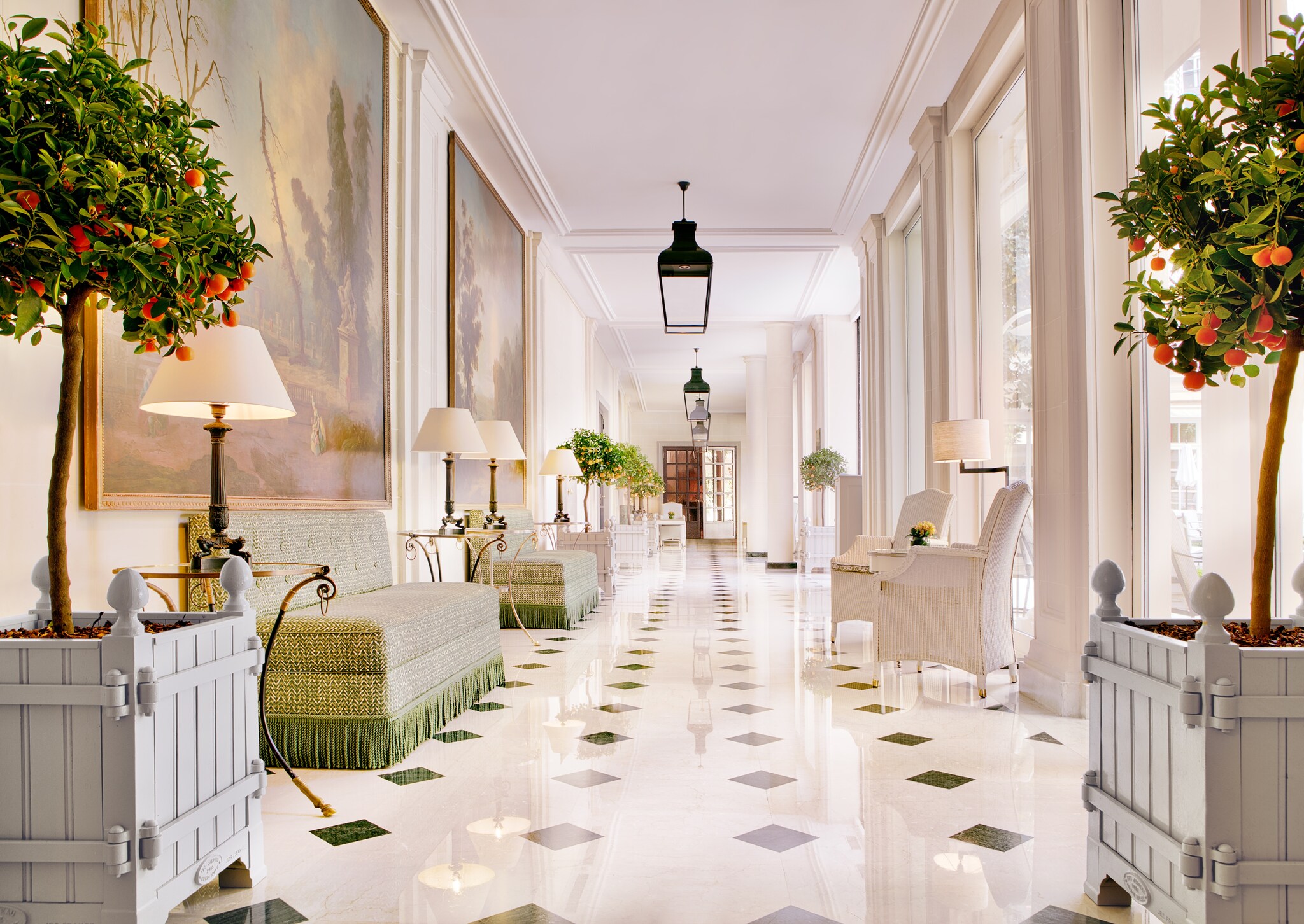 A long hallway is paved with gleaming white and black tiles and furnished with potted citrus trees and light-colored seating.