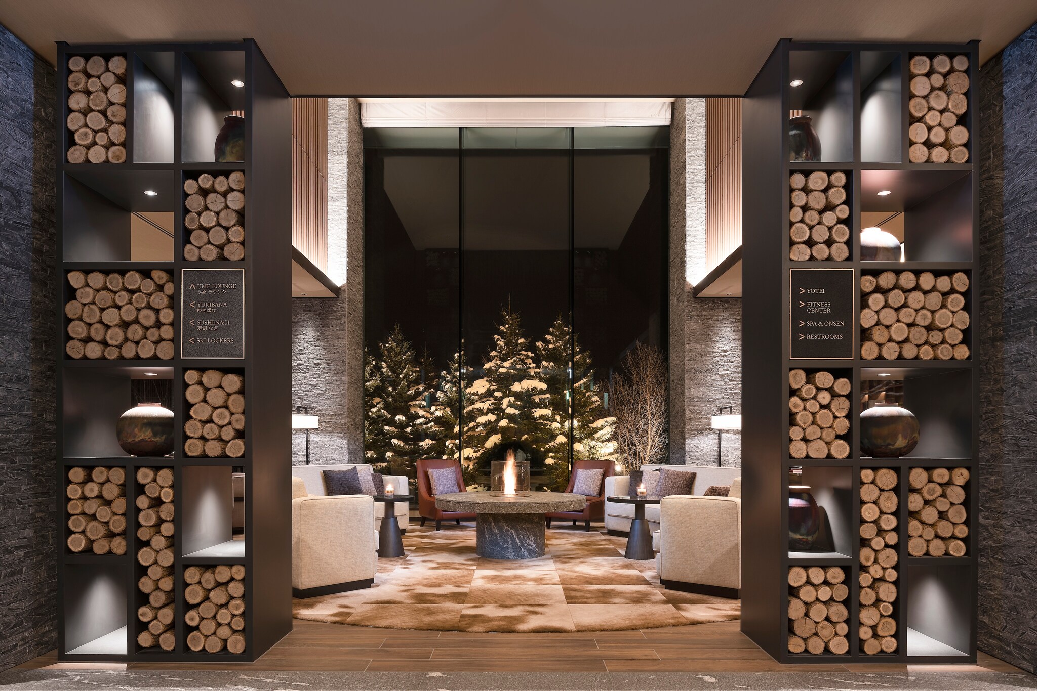 The lobby features high ceilings and tall bookshelves stacked with cut wood. Armchairs are arrayed around a circular table with a flame.