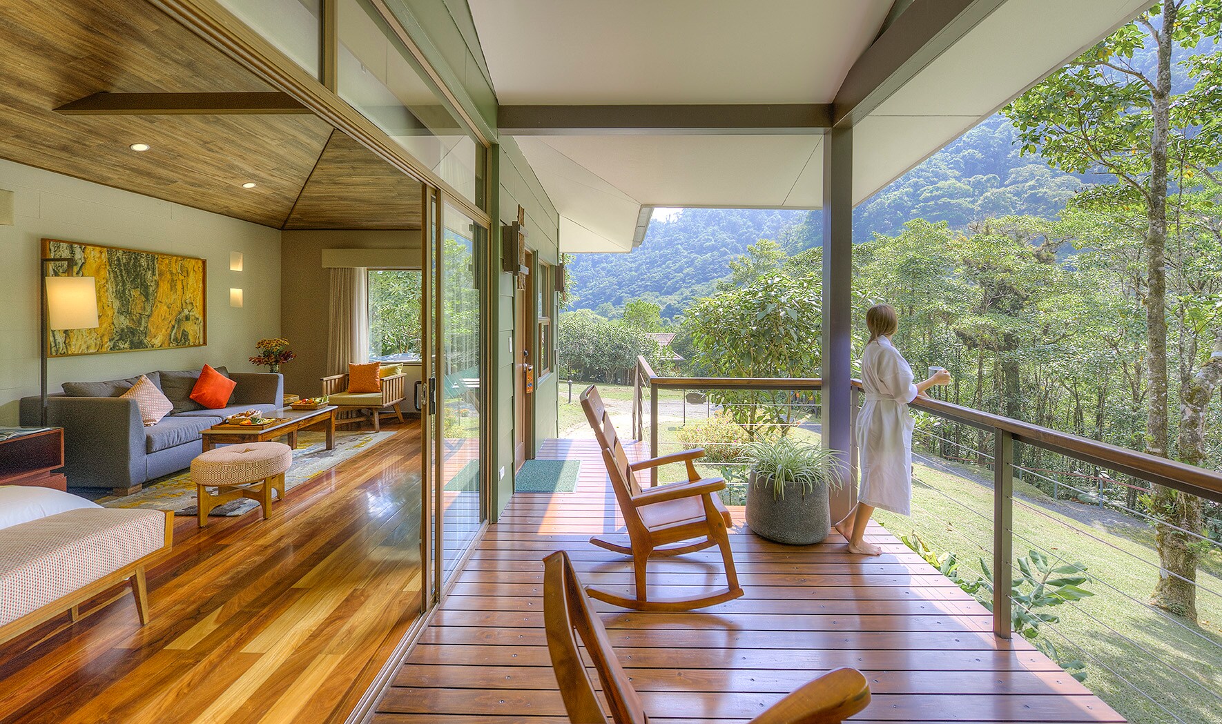 A woman in a white robe stands on a suite patio looking out at the jungle. Through open glass doors, an airy indoor living area is visible.