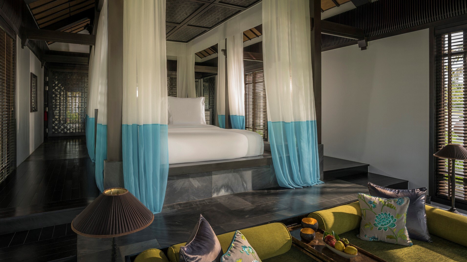 Interior of a one-bedroom villa. A canopy bed is draped with color-blocked white and blue curtains. Nearby, a green sofa is set into the floor.