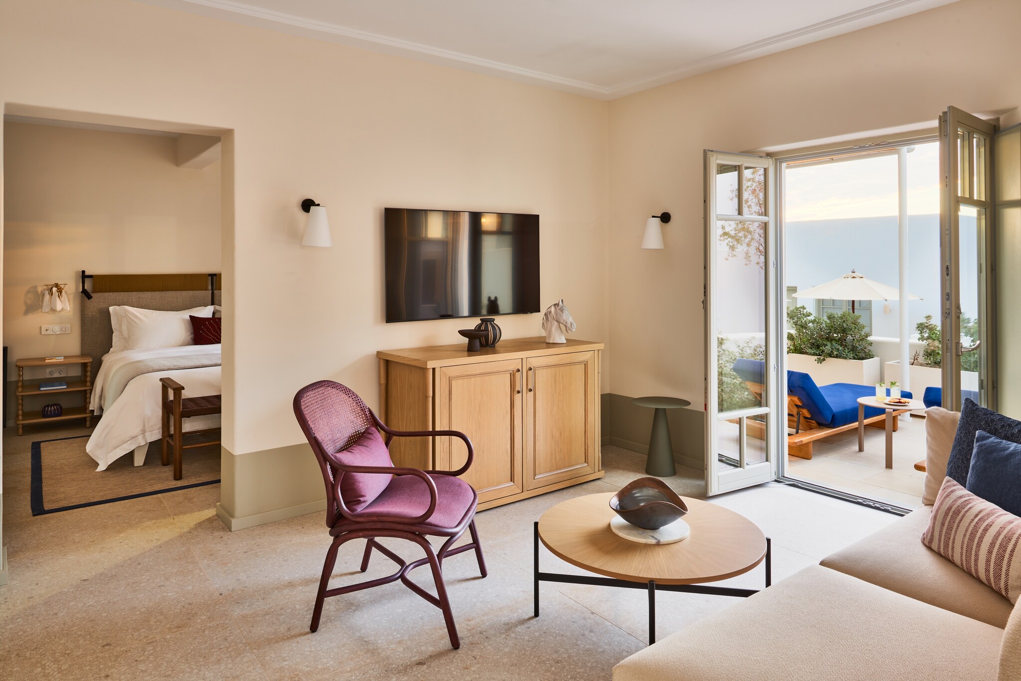 The living area of a guest room has a TV and a purple chair featuring a tan couch and opening out onto a small balcony.