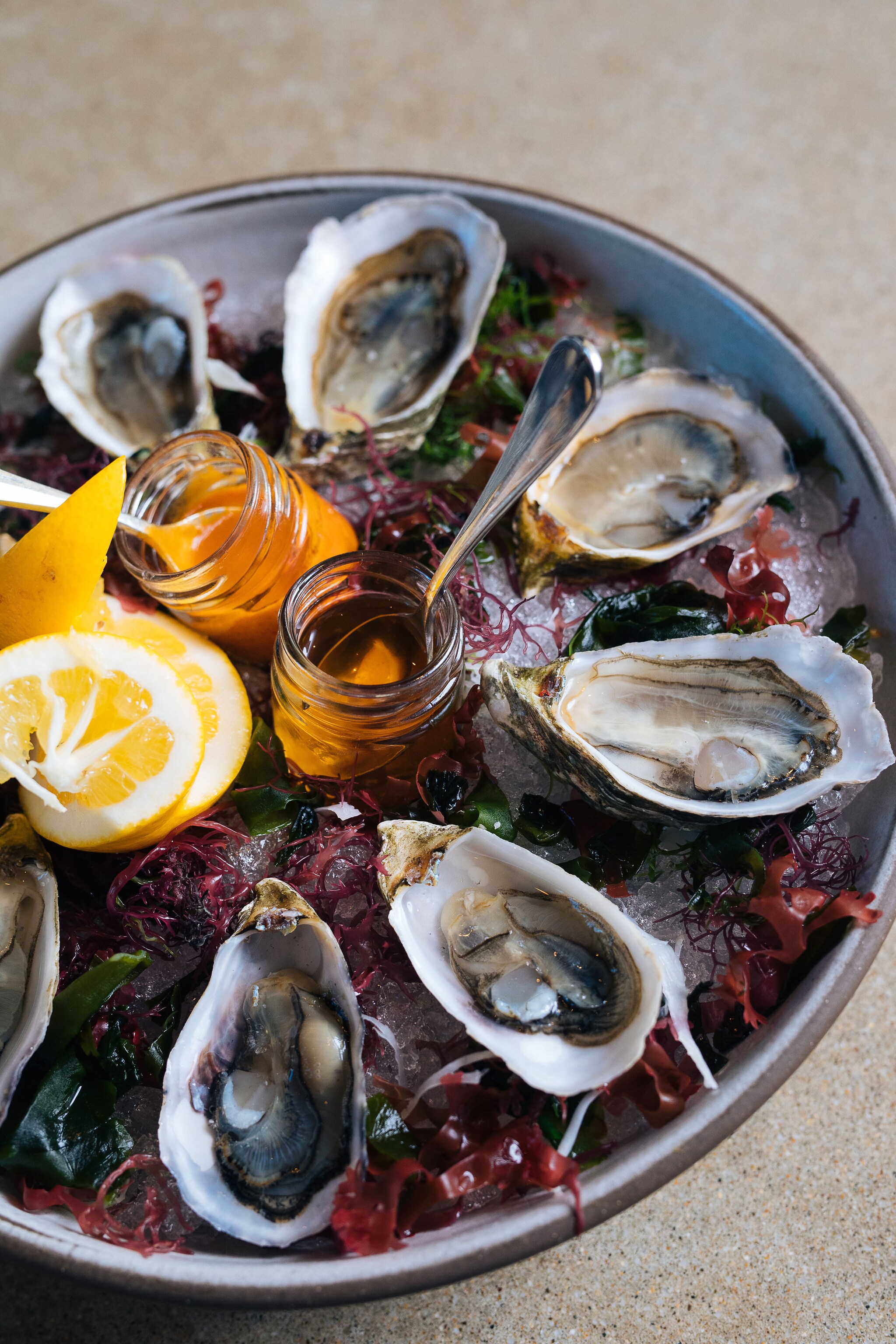 Close-up for an oyster tray, garnished with a curl of lemon, sprigs of purple seaweed, and accompanied by jars of amber sauce.