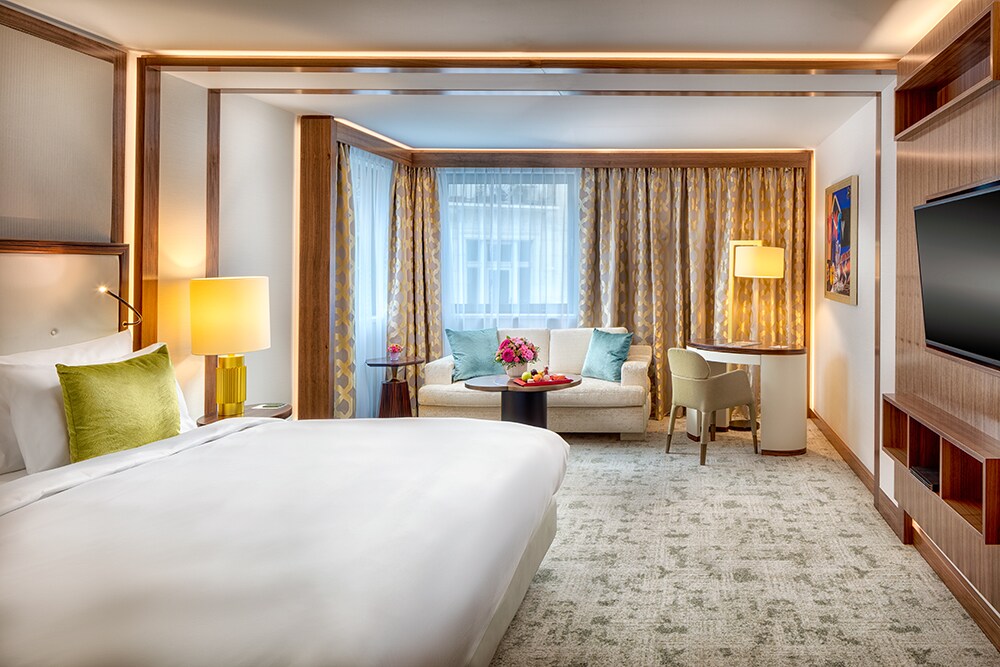 A large guest room has gray-green carpets and a white bed with a green accent pillow. A white loveseat and a round desk sit next to windows.