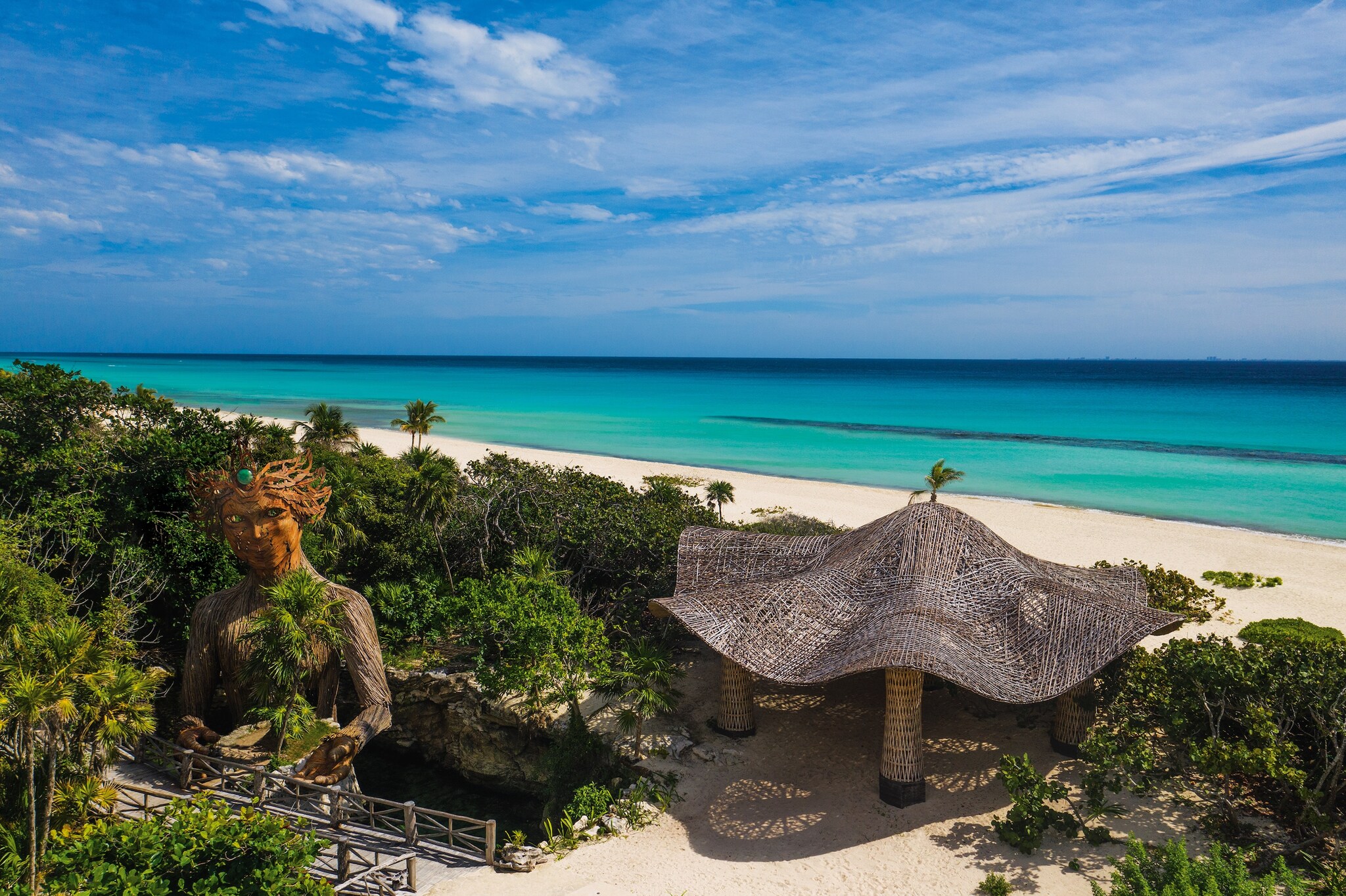 The beach is fronted by lush greenery, a rippling, mushroom-like gazebo, and a wooden statue of Aïa, a woman holding a small island.