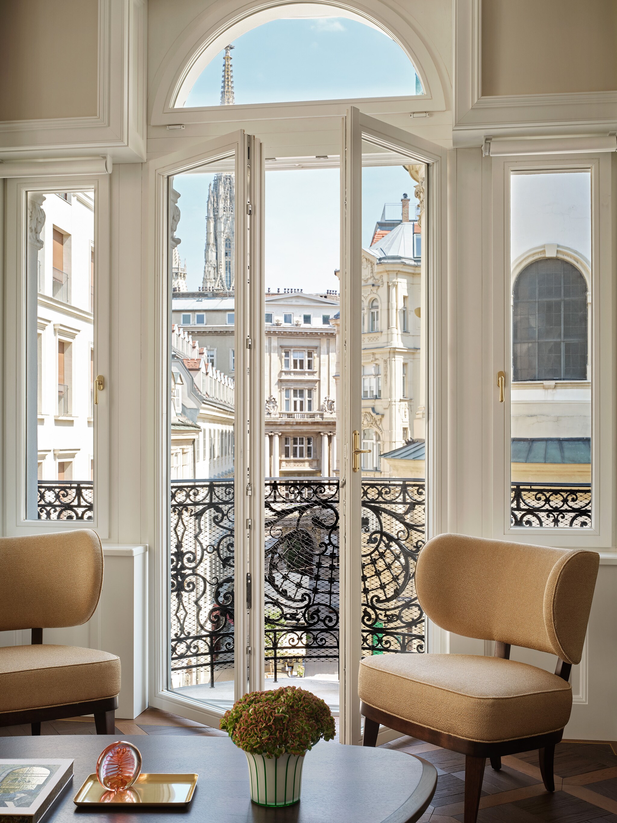 Past the glass doors and wrought-iron railing of a guest room balcony, the gray spire of St. Stephen’s Cathedral is visible.