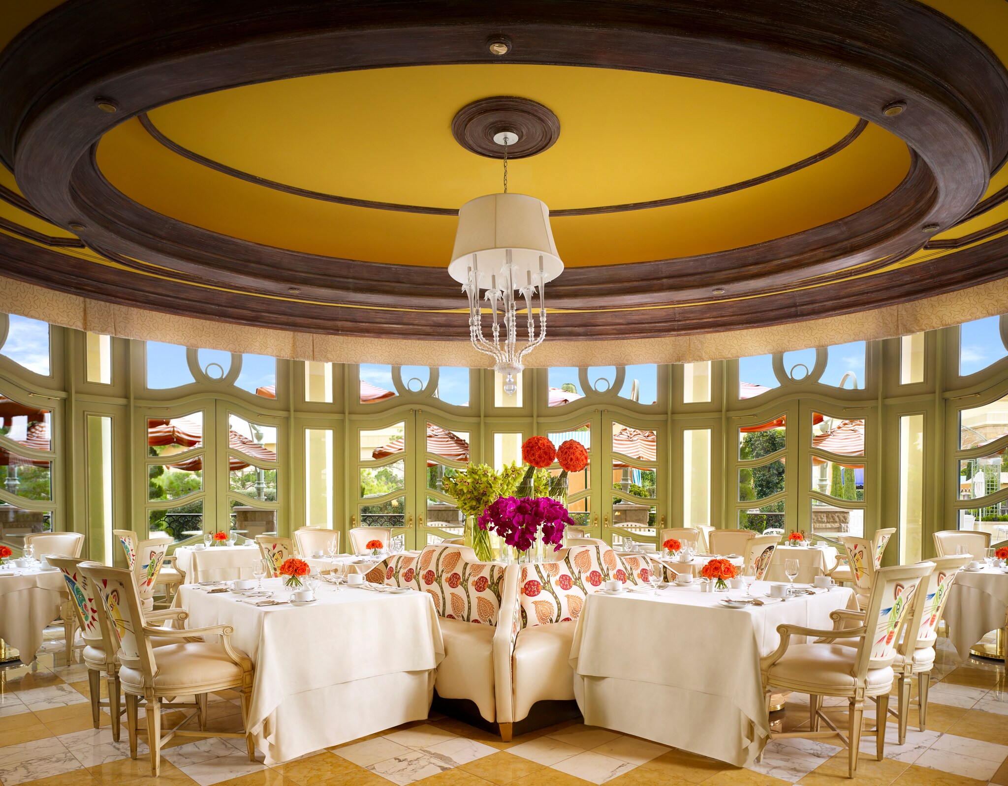 The Tableau Conservatory is decorated in white, cream, and gold, with floral details and green window frames looking out onto a lush patio.