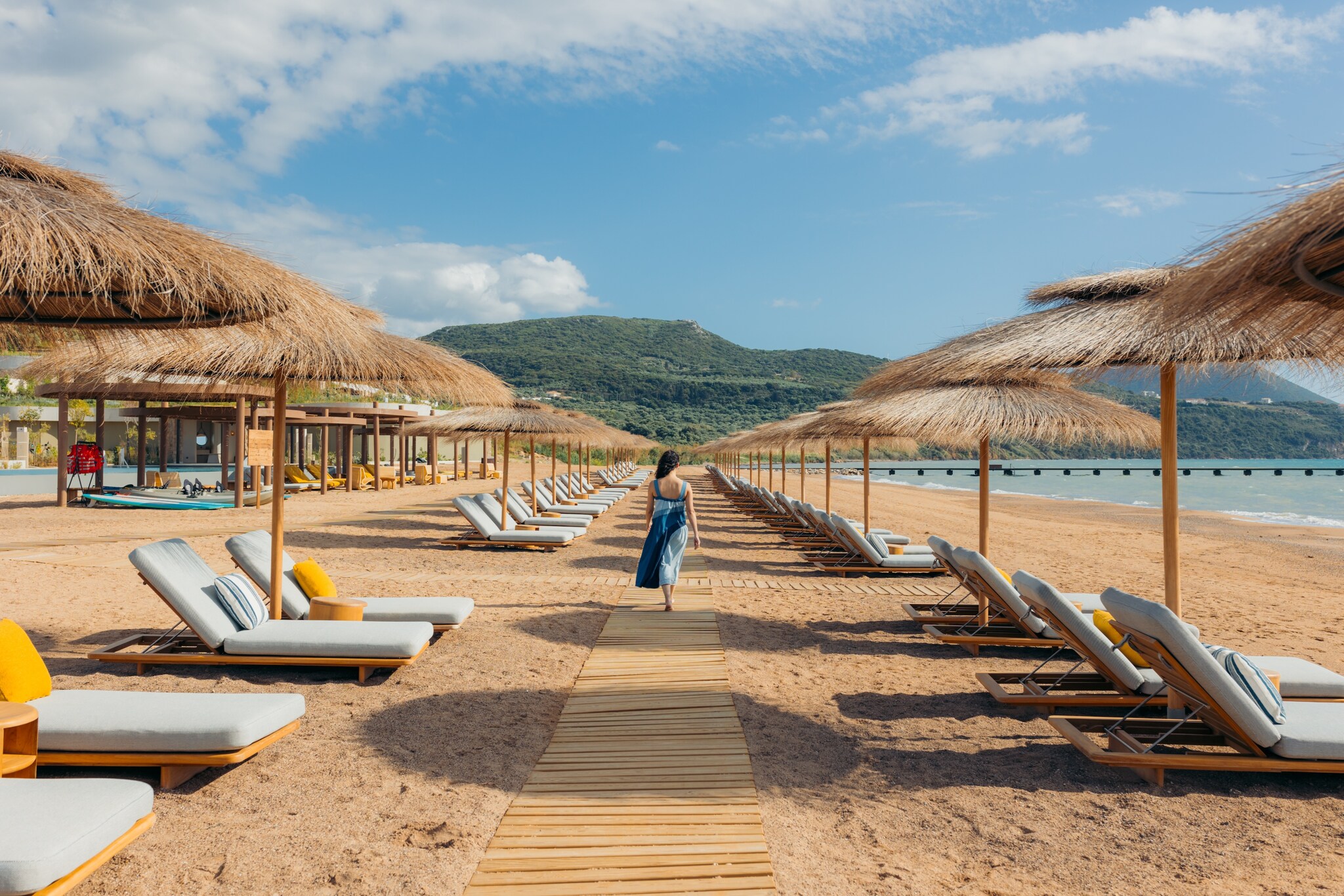A white woman with long, dark hair walks away from the viewer down a boardwalk set onto the beach between rows of sun beds and umbrellas.