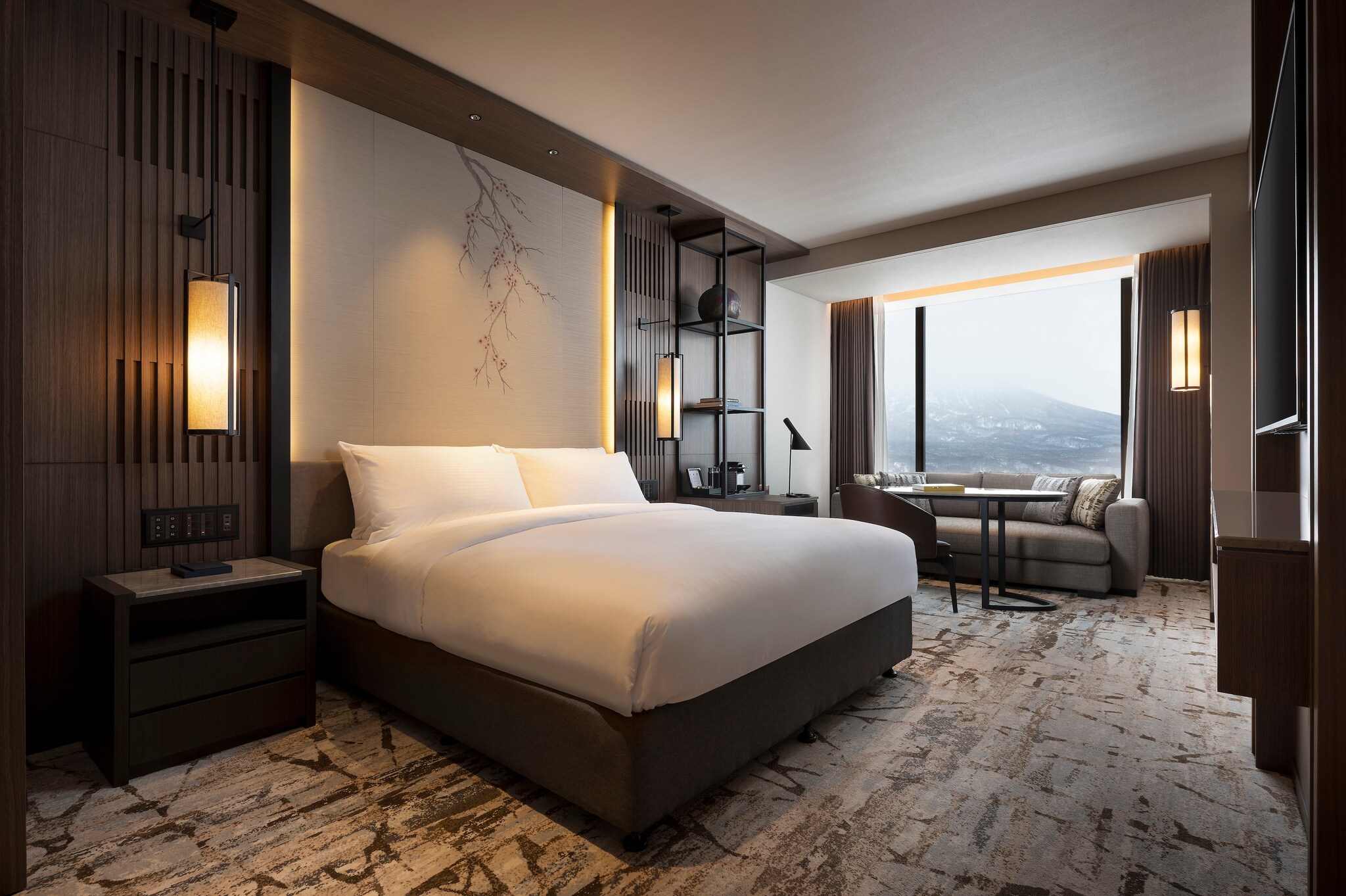 A guest room has a bed with a tall headboard with a minimalist floral design. On the far wall, a small gray couch rests under tall windows.