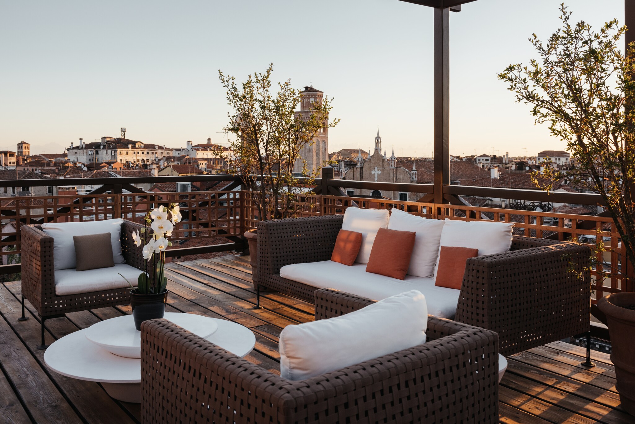 On the roof terrace, brown wicker seating with white cushions is accented with potted trees. Beyond which the city stretches all around.