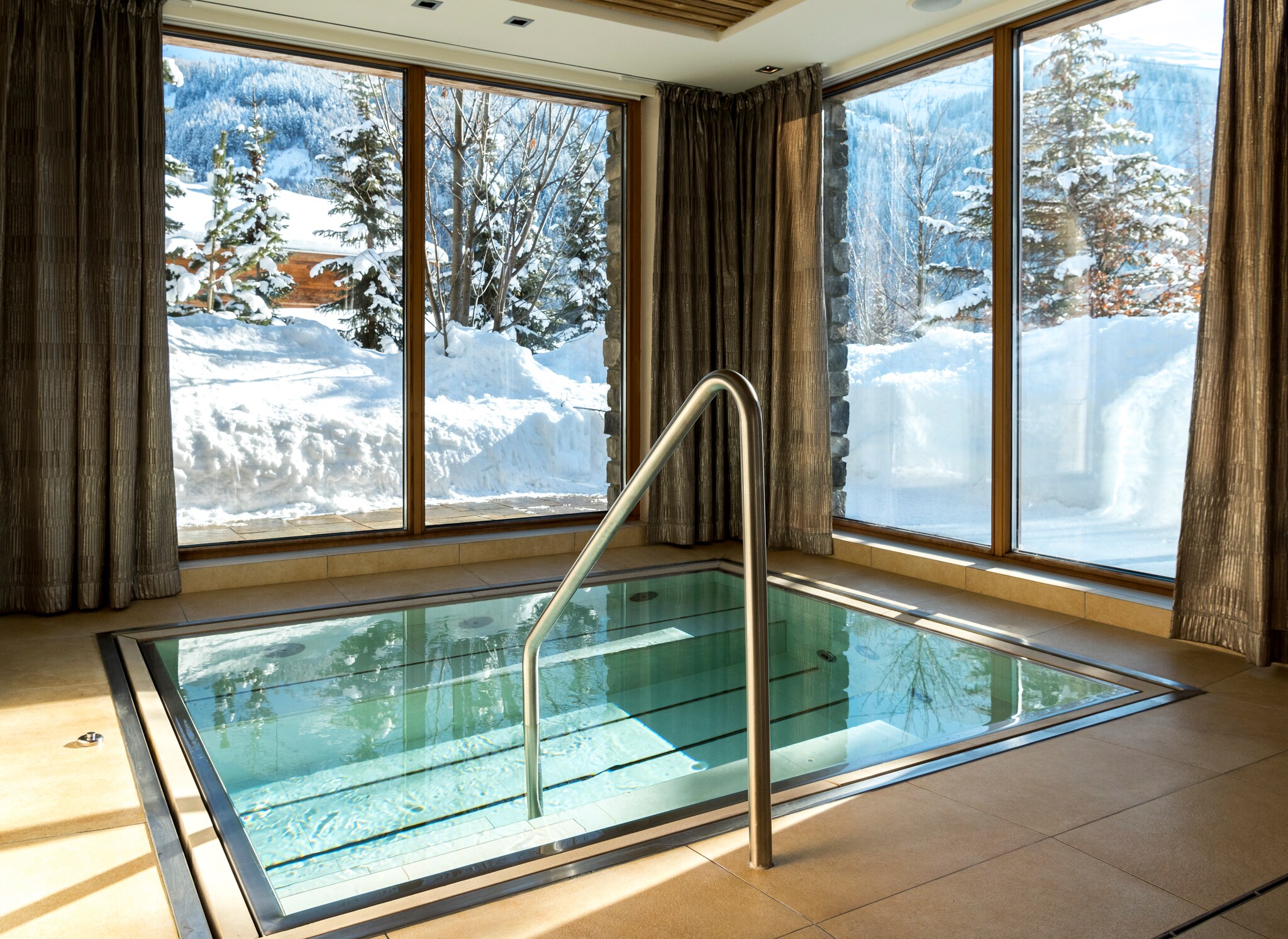 A square whirlpool in a pool room is surrounded by long curtains and tall windows revealing deep snow.