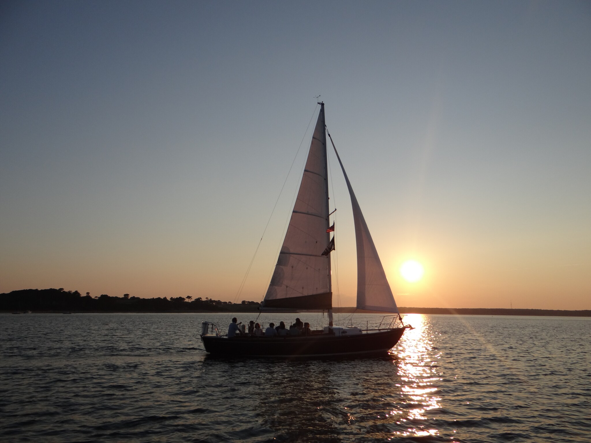 A sailboat with people crowded together on the deck is silhouetted on the water in front of a sunset.