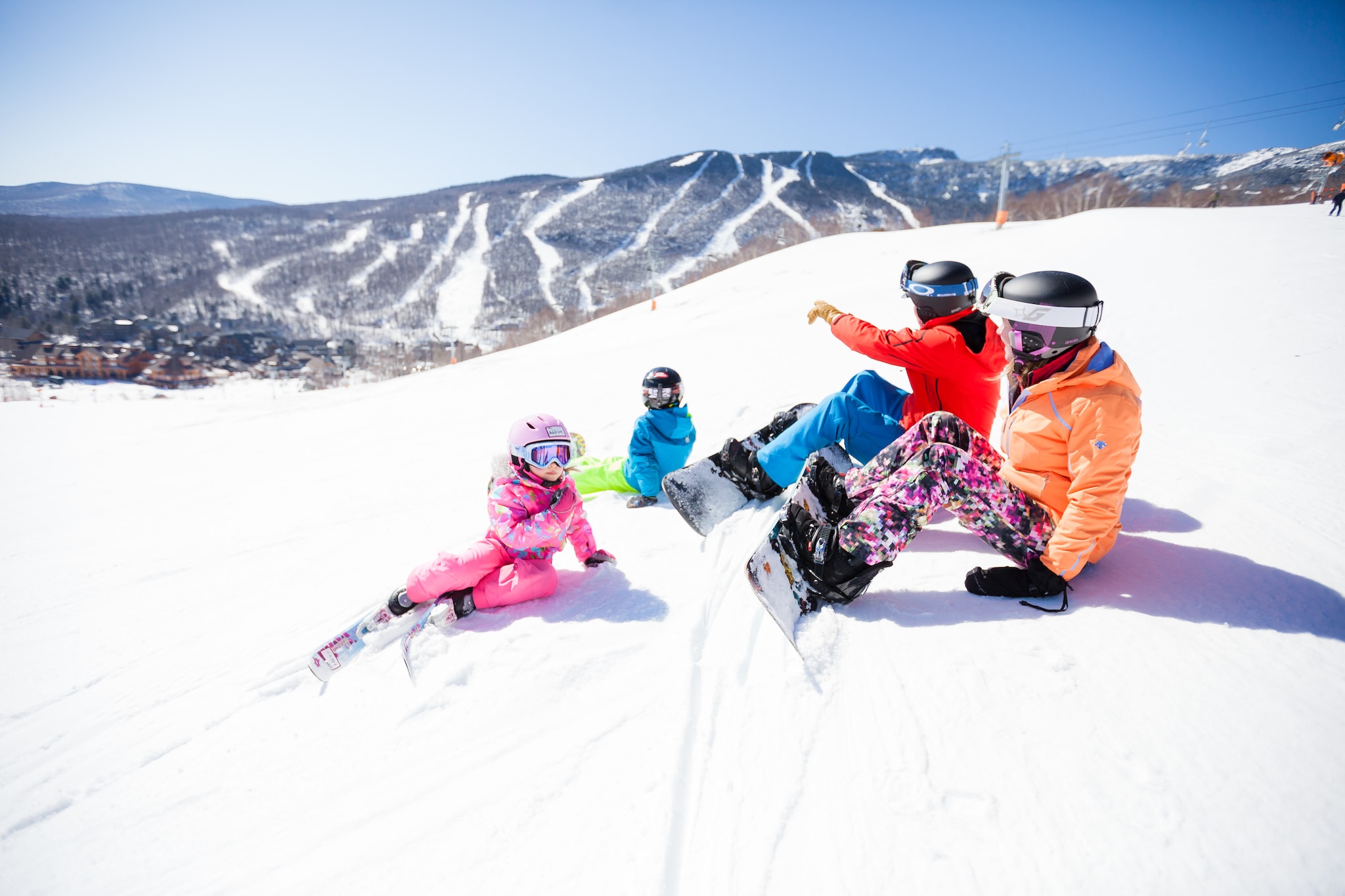A family in brightly-colored snow gear sits and chats on a ski slope. The parents are clipped into snowboards and the children wear skis.