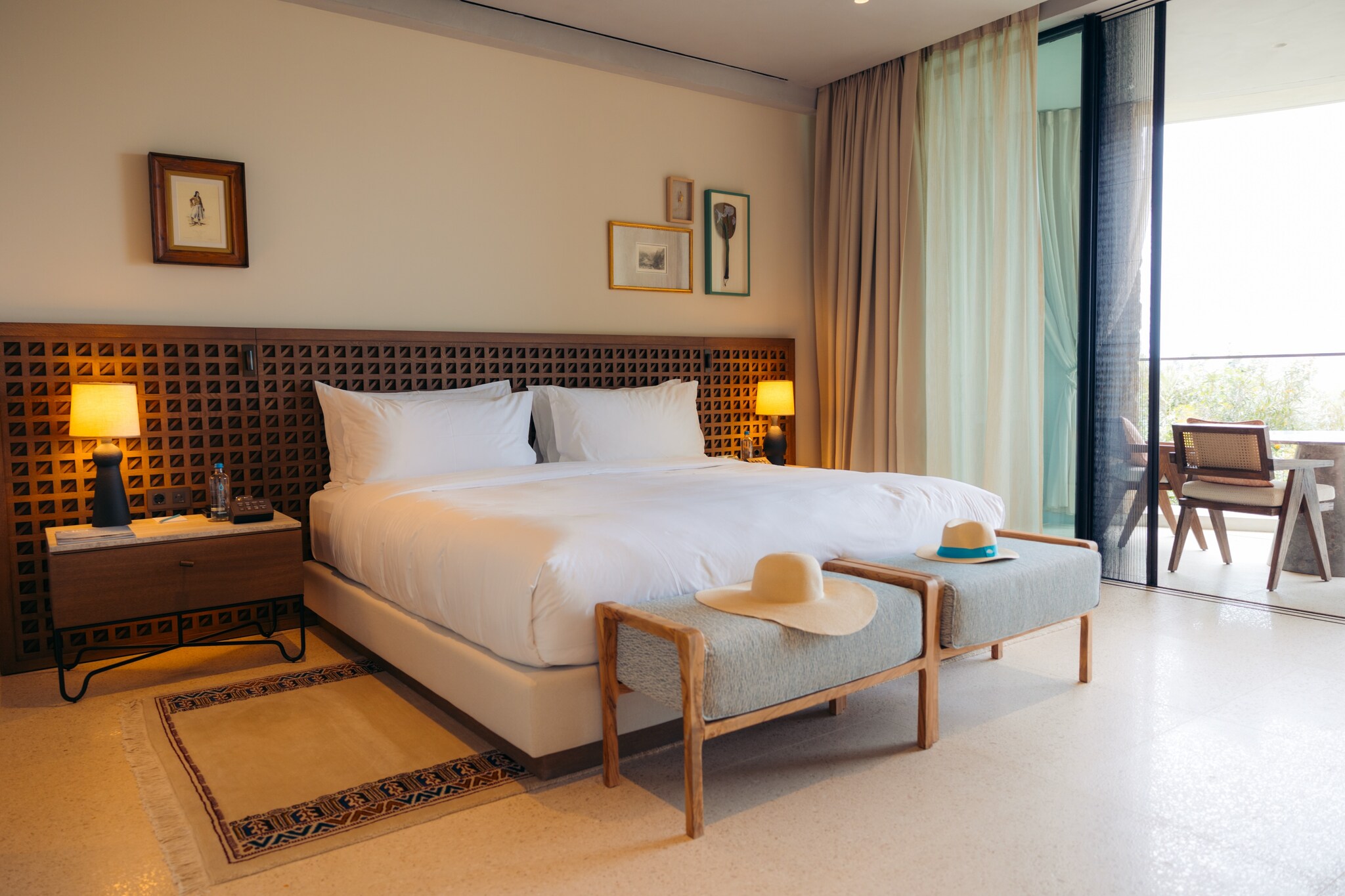 In a guest room, a wide bed has a dark wood grid at its head and two small benches at its foot, each with a sun hat. Beyond is a sunny patio.