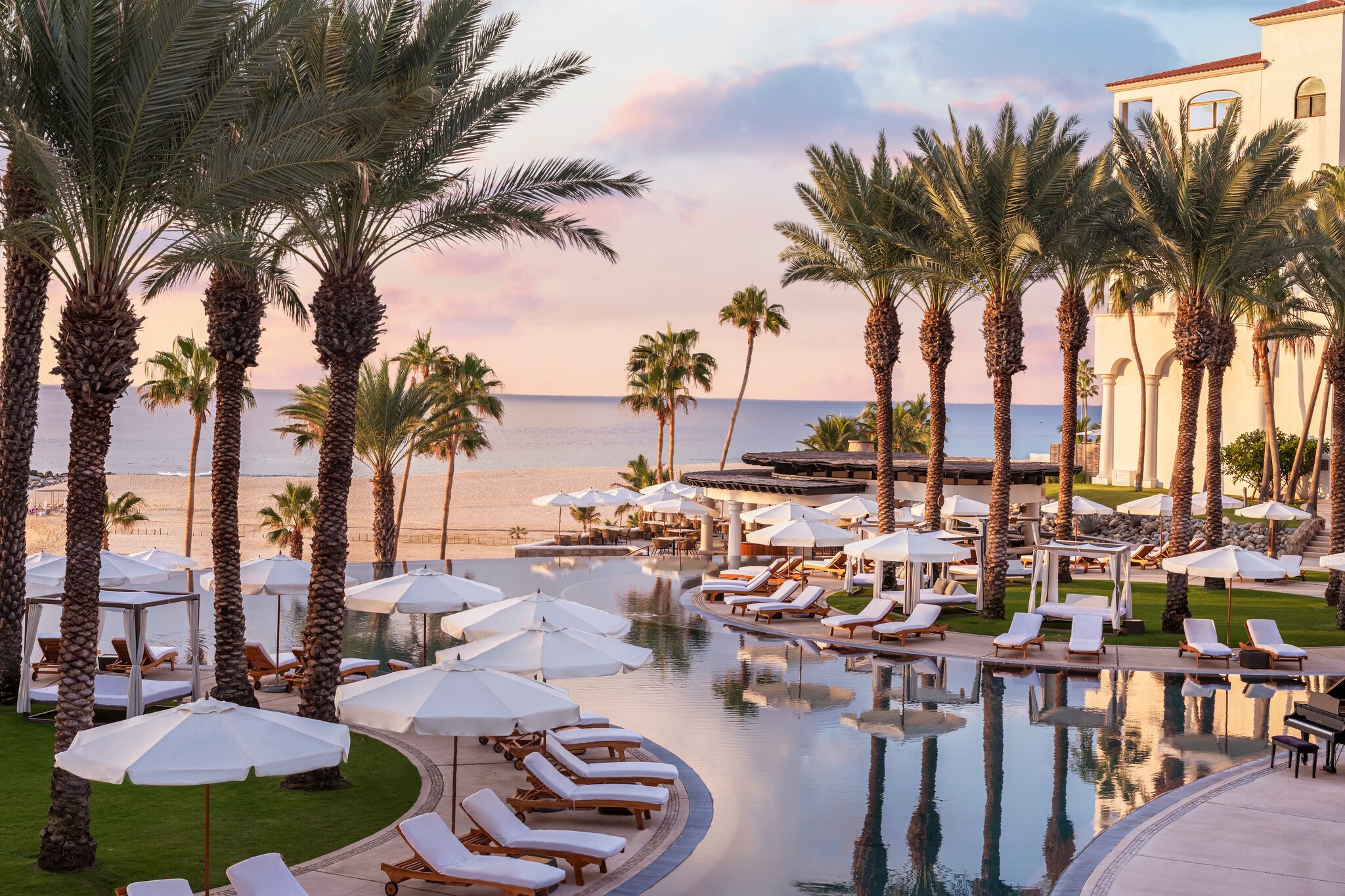 The pool at Hilton Los Cabos runs between curved decks lined with sun loungers, umbrellas, and palm trees near the beach.