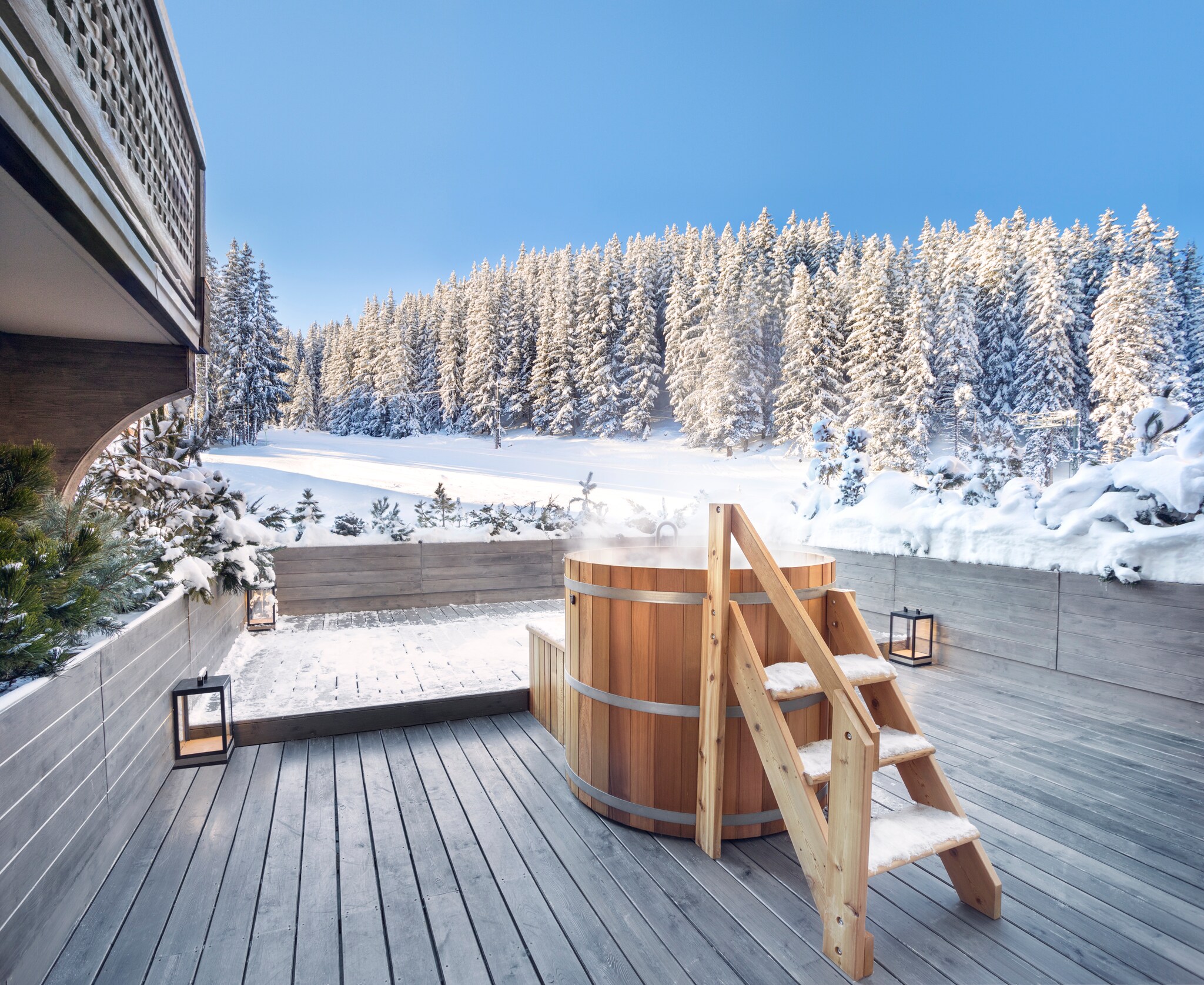 On the deck of a Chambre Ski Piste suite, a small set of wooden stairs leads into a steaming wooden hot tub. Beyond is thick snow and trees.
