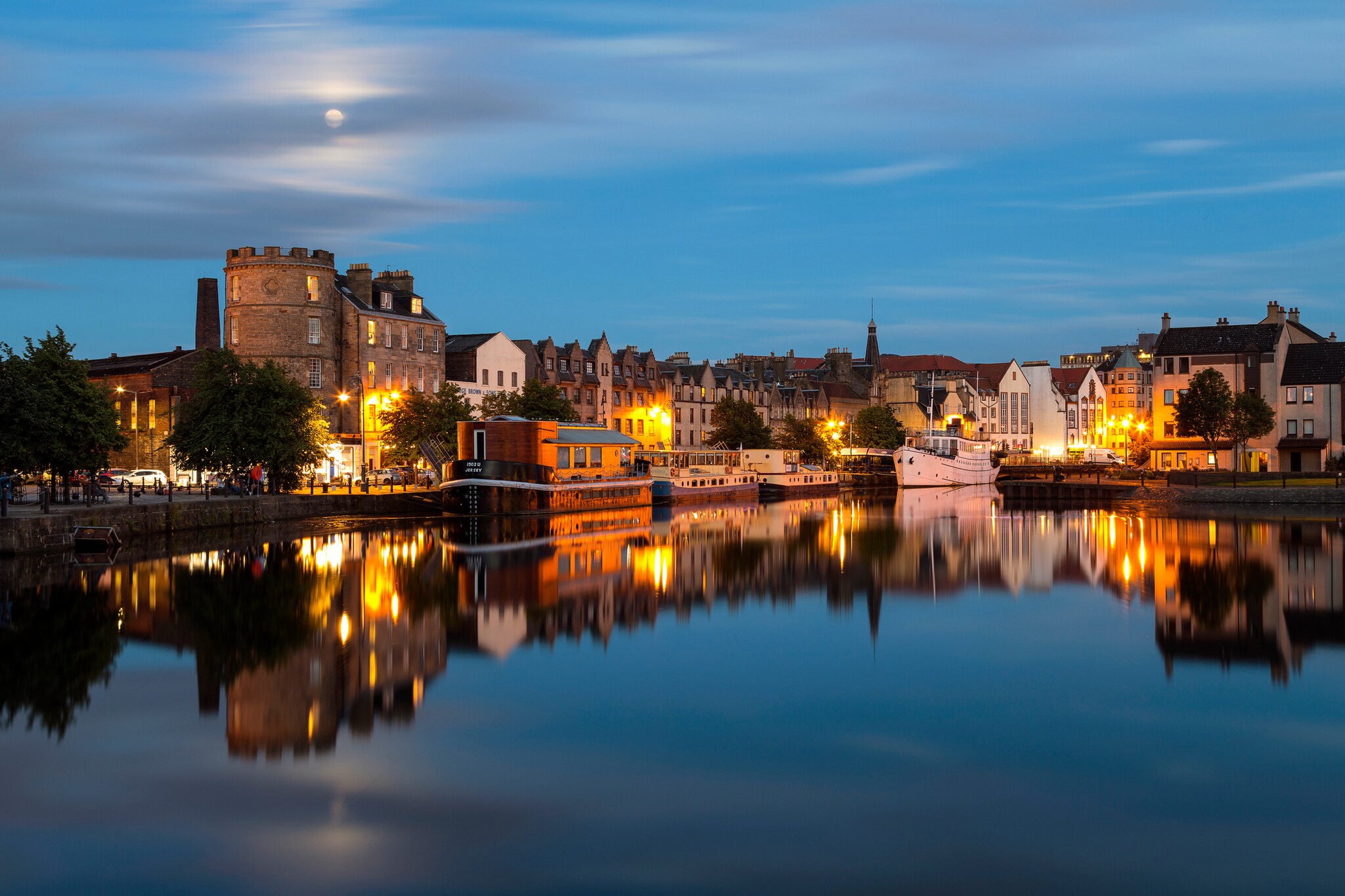 Evening view across the Water of Leith at the Signal Tower topped with battlements. Stout boats and streetlights are reflected in the water.