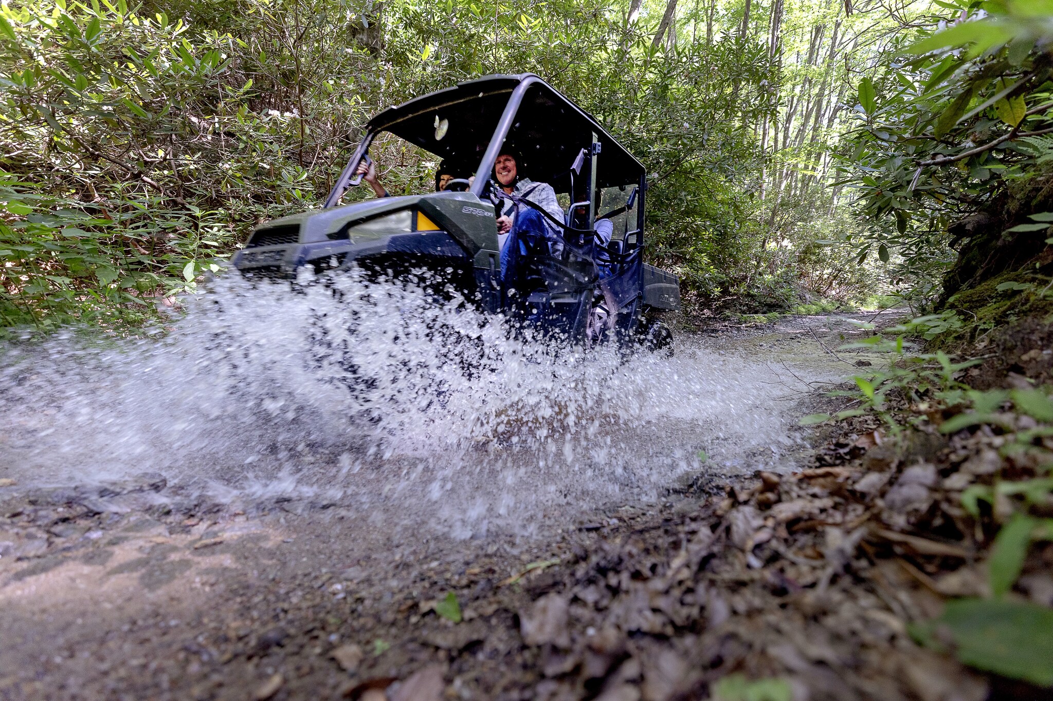 Guests in helmets driving a Recreational Terrain Vehicle down a forest path, splashing water from a puddle towards the camera.