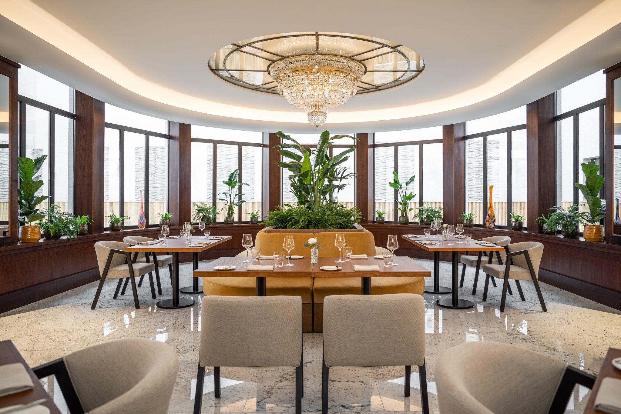 The Alcron Restaurant dining room is circular, with a central bank of ochre bouches built around a large potted plant under a chandelier. 