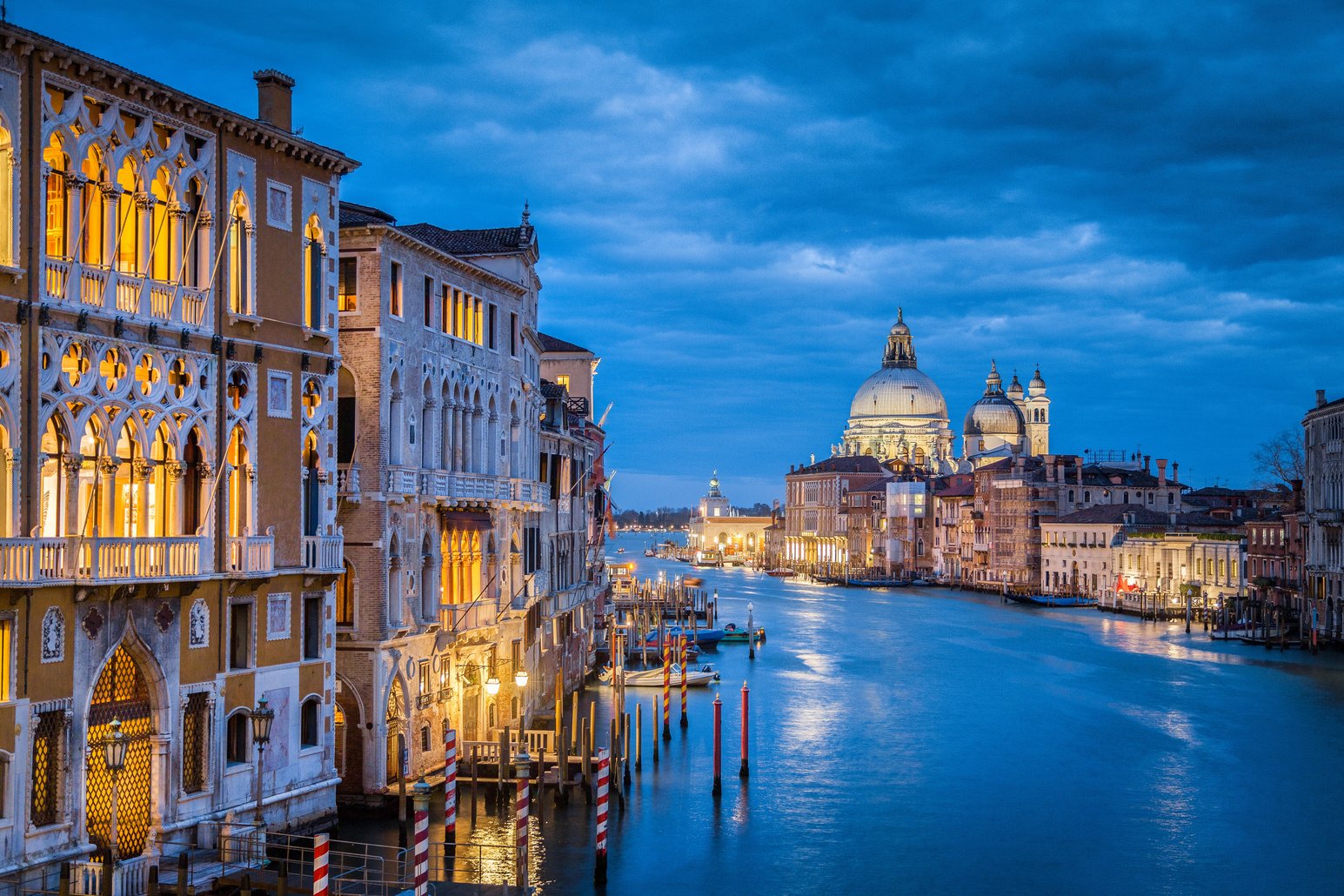 Early-evening view past buildings with glowing windows over the canal, looking towards the dome of Baslica Santa Maria della Salute.