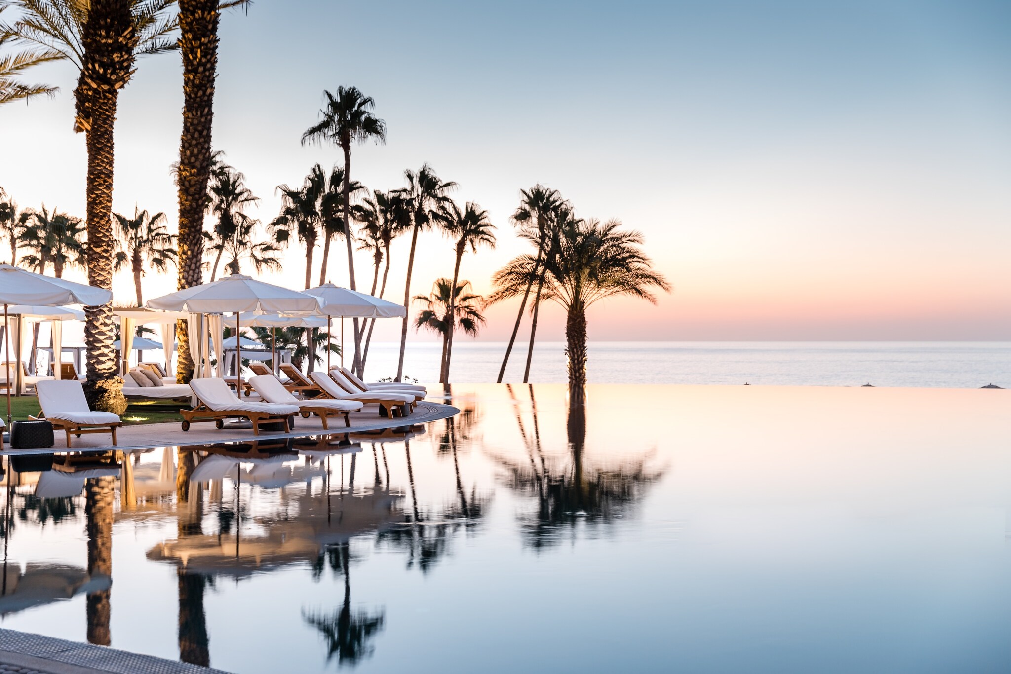 An infinity pool reflects the ombre sunset sky, palm trees, as well as the sun loungers and umbrellas on the deck.