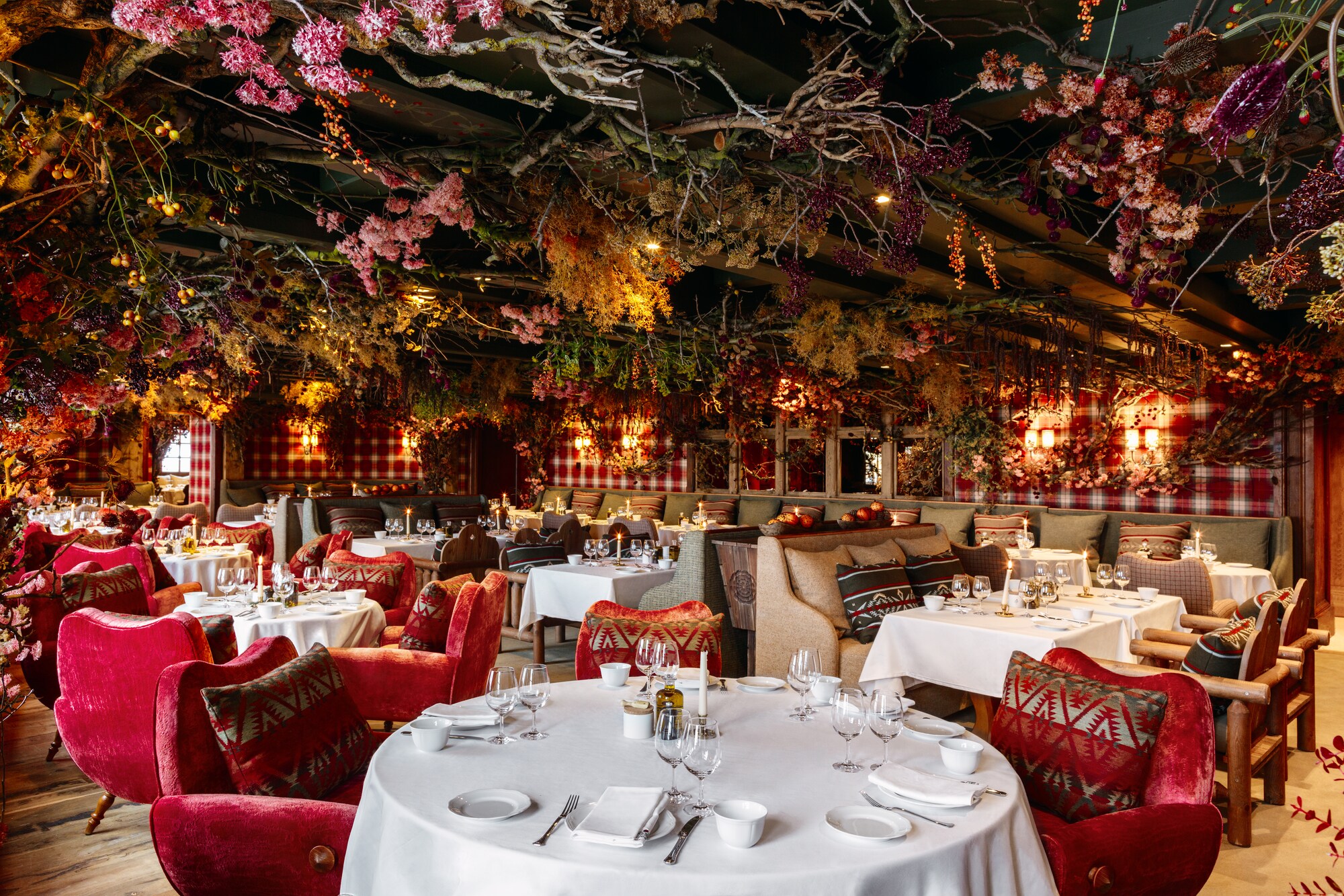 The ceiling of a dining room is lush with flowers and colorful branches. Underneath, red velvet chairs are stationed around round tables.