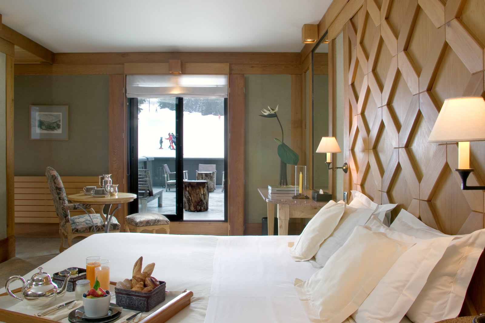 A tray of pastries, fruit, tea, and juice is laid on a large white bed under a latticed headboard. Beyond, glass doors open to a patio.