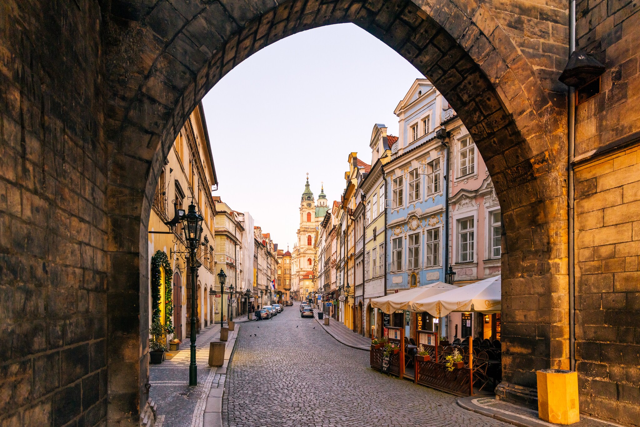 Looking through an arch down a cobblestone street in Prague’s Old Town. The street is lined with pastel buildings with ornate white trim.