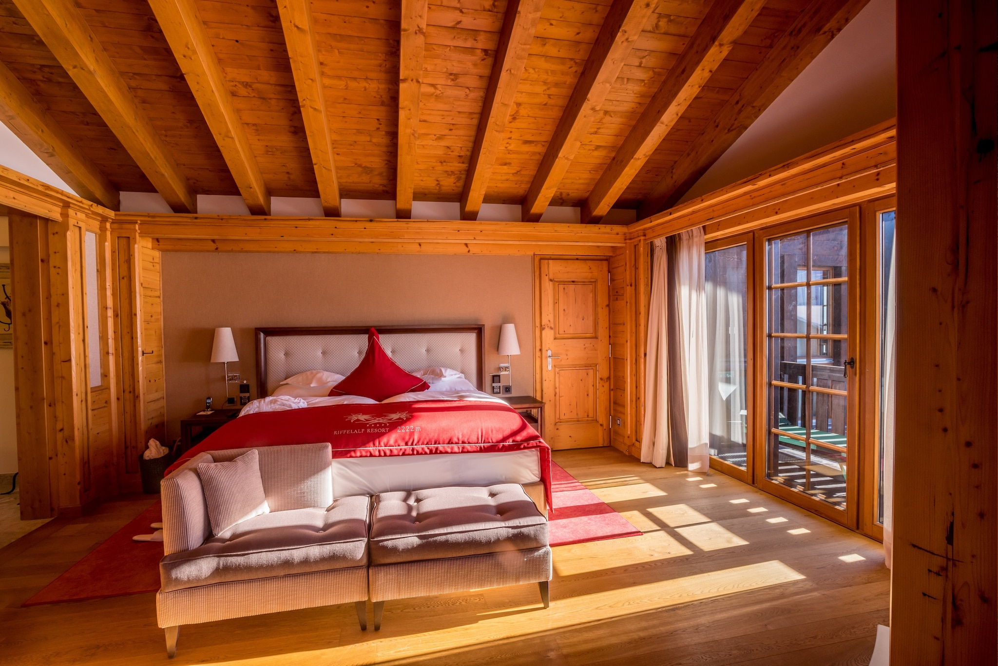 The sleeping room of a suite is paneled with knotted wood. A large white bed is accented with a red duvet cover and red triangular pillow.