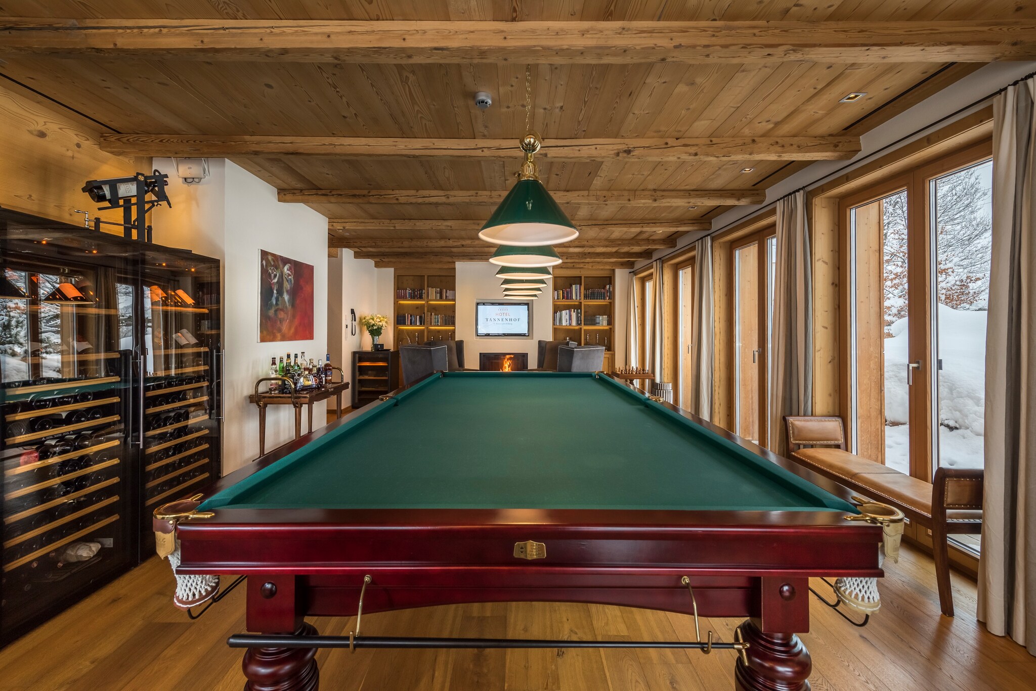 View over a billiards table. One nearby wall is lined with floor-to-ceiling windows and another is lined with cabinets of wine.