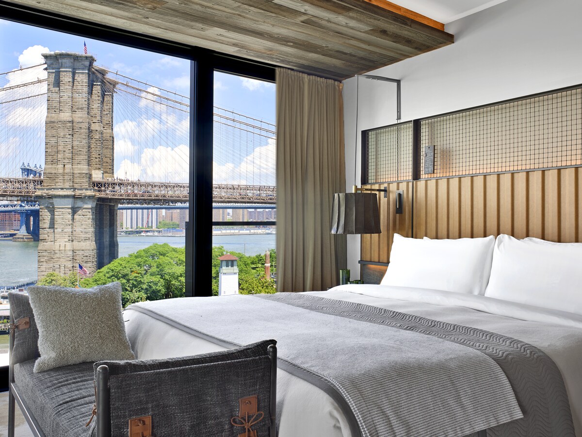 A Bridge King Guestroom features a large bed with a sleek headboard. The Brooklyn Bridge is prominent outside the floor-to-ceiling windows.