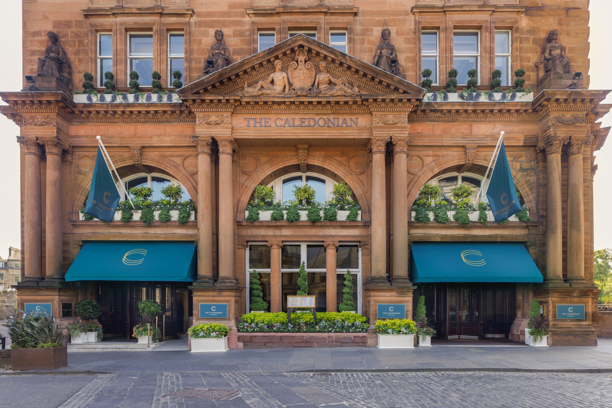 Exterior of The Caledonian Edinburgh, which has a neoclassical design in red stone, and pots of plants and spiral trees lining the windows.