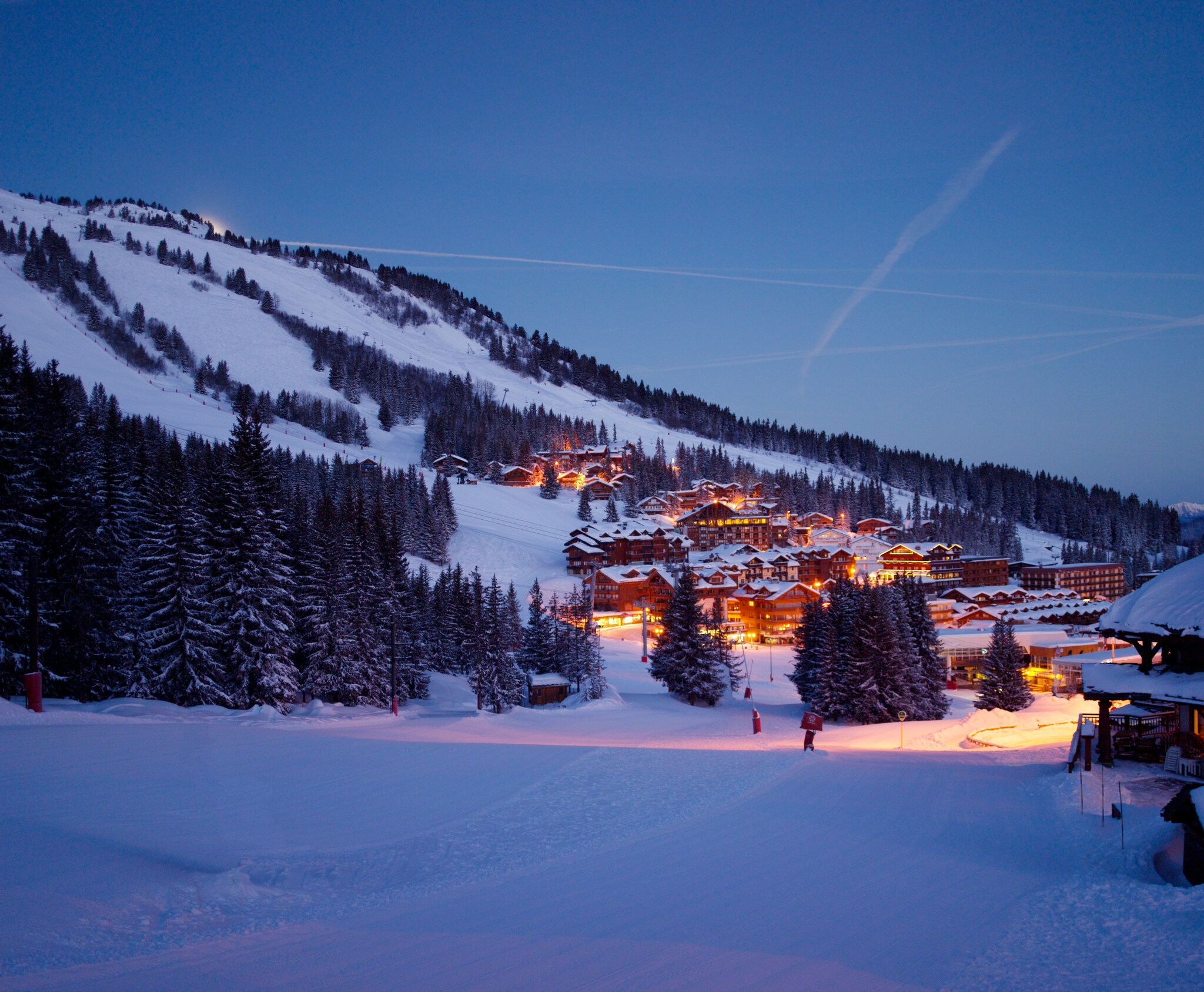 Night view of Courchevel village, with rows of buildings lit up in the valley between snowy, forested mountains.