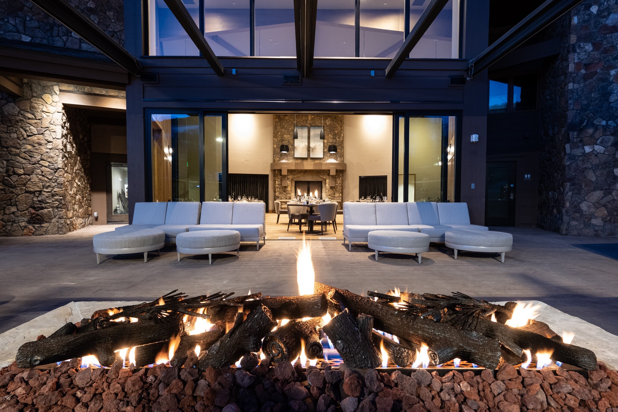 Outdoors, a lit fire pit sits between the viewer and a set of white furniture. Wide glass doors are open to reveal a brightly-lit room.