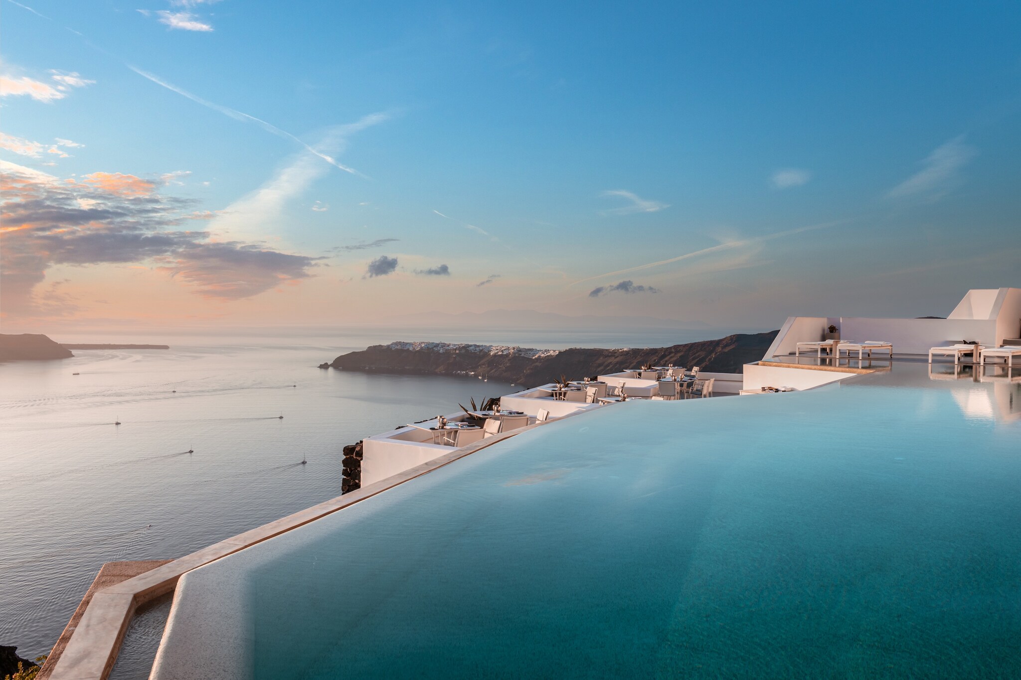 An infinity pool stretches out to the edge of a white balcony and overlooks the rocky island and the smooth water of the Aegean Sea.