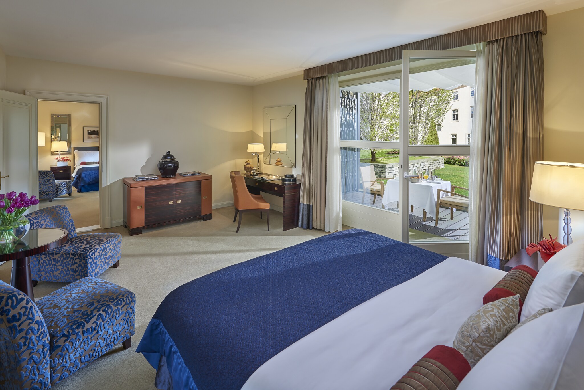 A sleeping room is arrayed with a bed and blue cut-velvet chairs. Glass doors open onto a patio and another doorway opens to another bedroom.