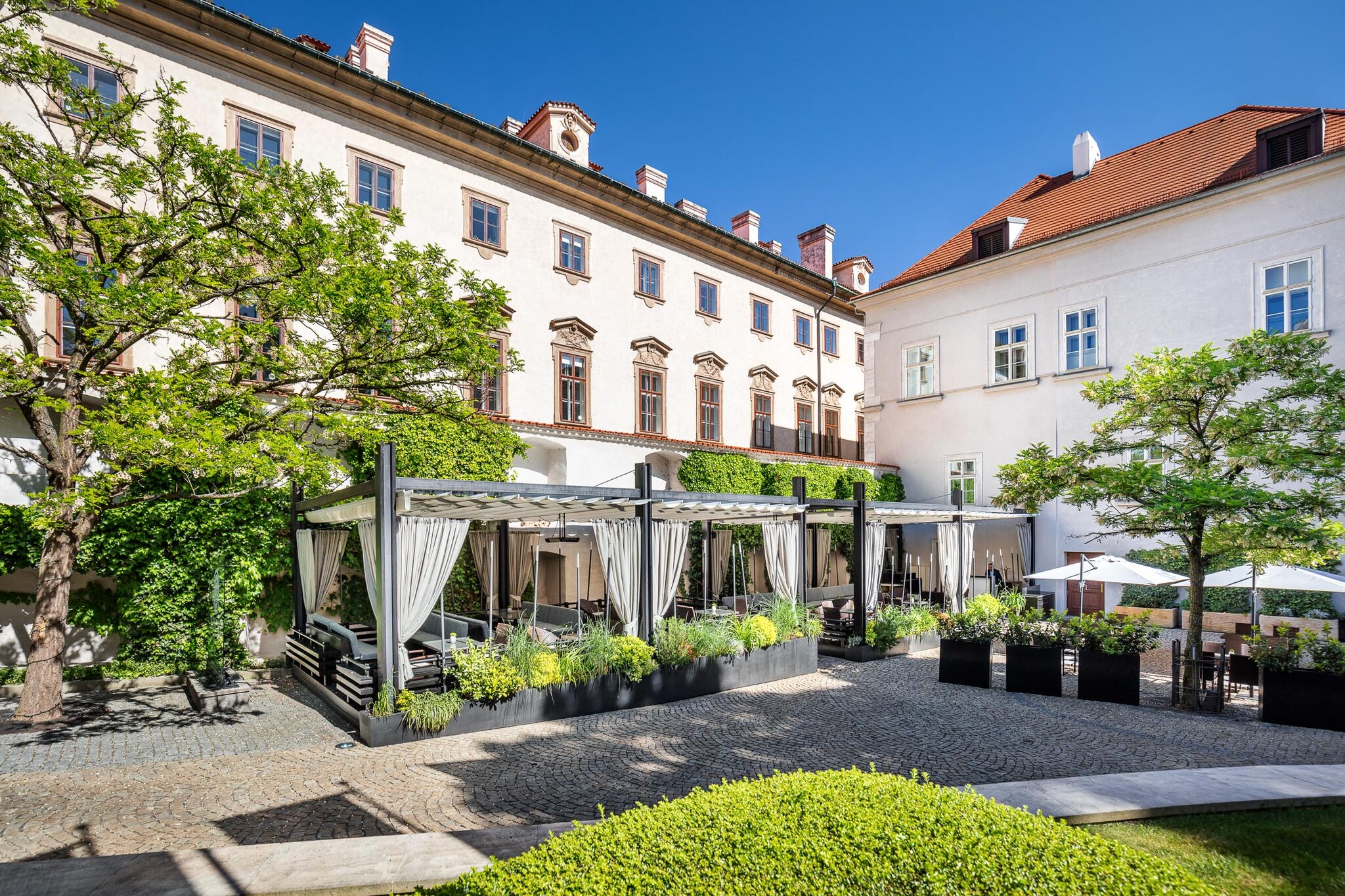 The hotel courtyard is filled with trees and potted plants, and dark couches are clustered under pergolas with cream curtains.