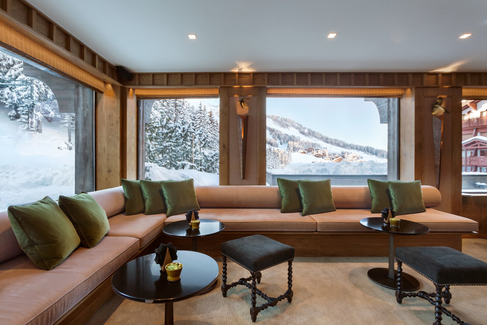 A large pink L-shaped couch is fitted into a corner against huge windows overlooking the slopes. Small black tables hold bowls of snacks.